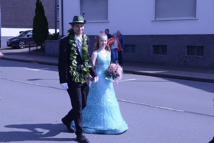 Ein junger Mann in Uniform und Hut führt eine junge Frau in einem hellblauen Abendkleid an der Hand. Im Hintergrund stehen Zuschauer und ein graues Gebäude.