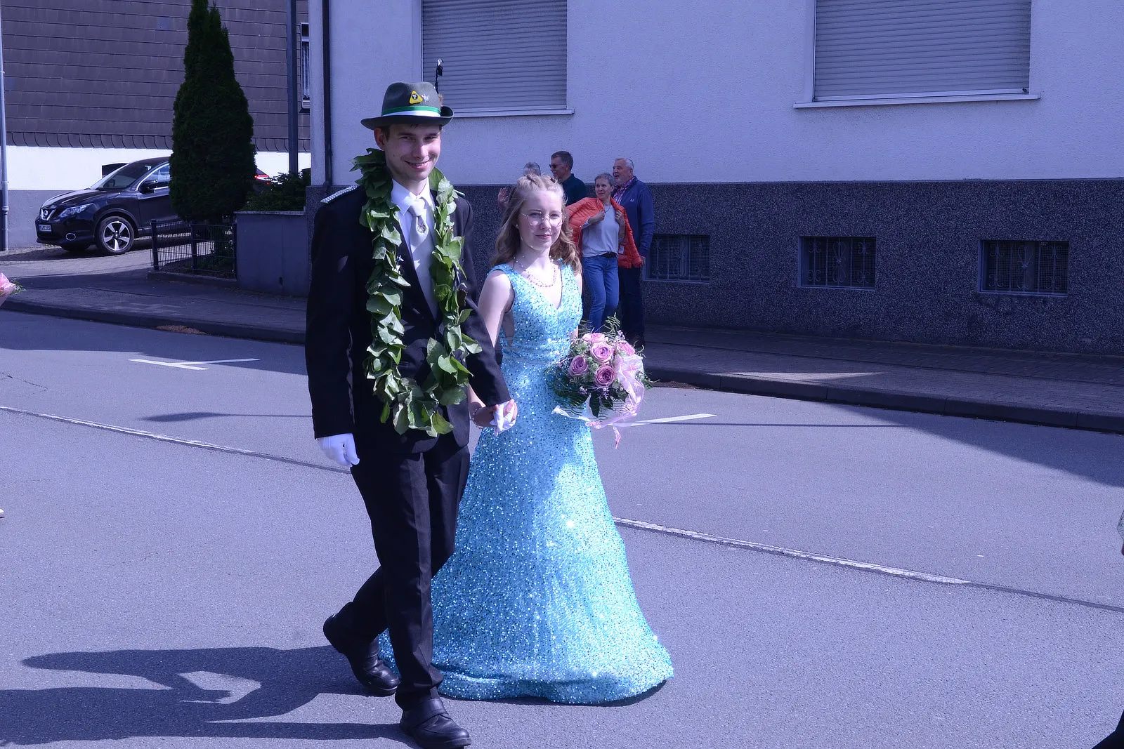 Ein junger Mann in Uniform und Hut führt eine junge Frau in einem hellblauen Abendkleid an der Hand. Im Hintergrund stehen Zuschauer und ein graues Gebäude.