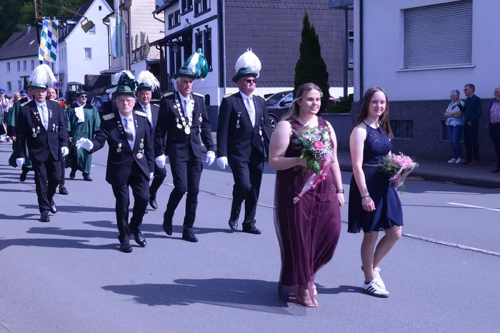 Festumzug mit festlich gekleideten Männern in Uniformen und zwei jungen Frauen mit Blumensträußen.