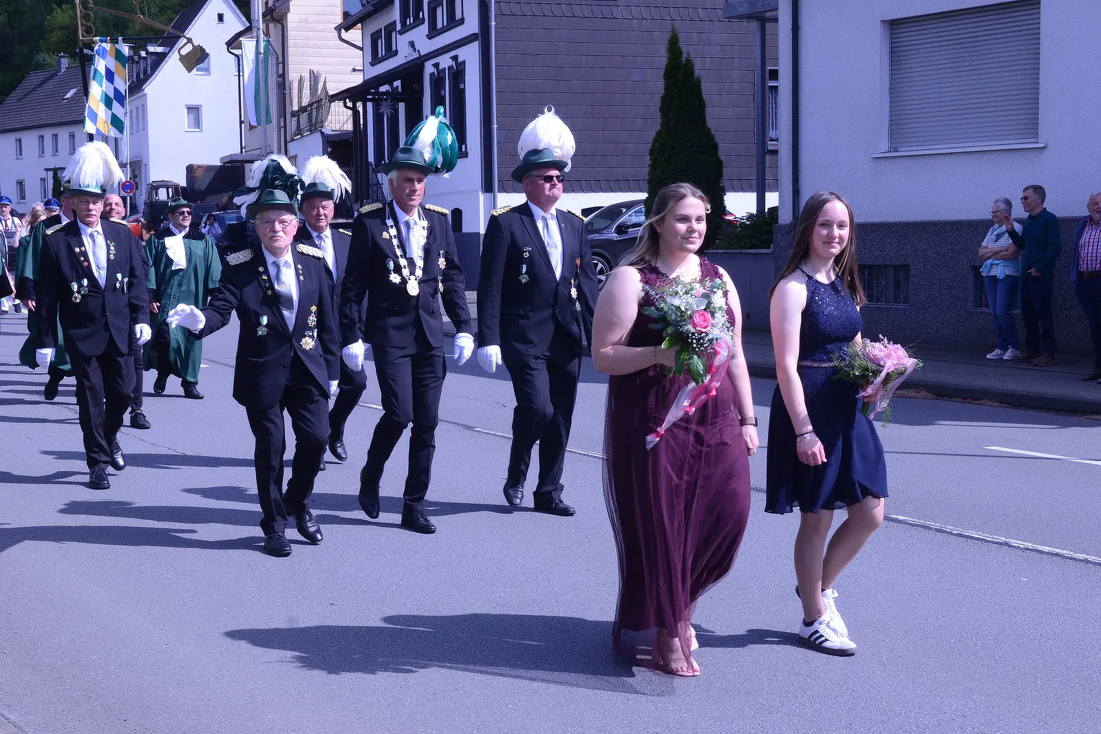Festumzug mit festlich gekleideten Männern in Uniformen und zwei jungen Frauen mit Blumensträußen.
