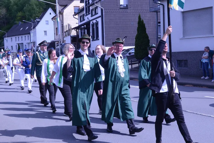 Festumzug mit Musikern in Uniformen und Personen in traditioneller Kleidung, darunter grüne Mäntel und Hüte, zieht an Häusern vorbei.