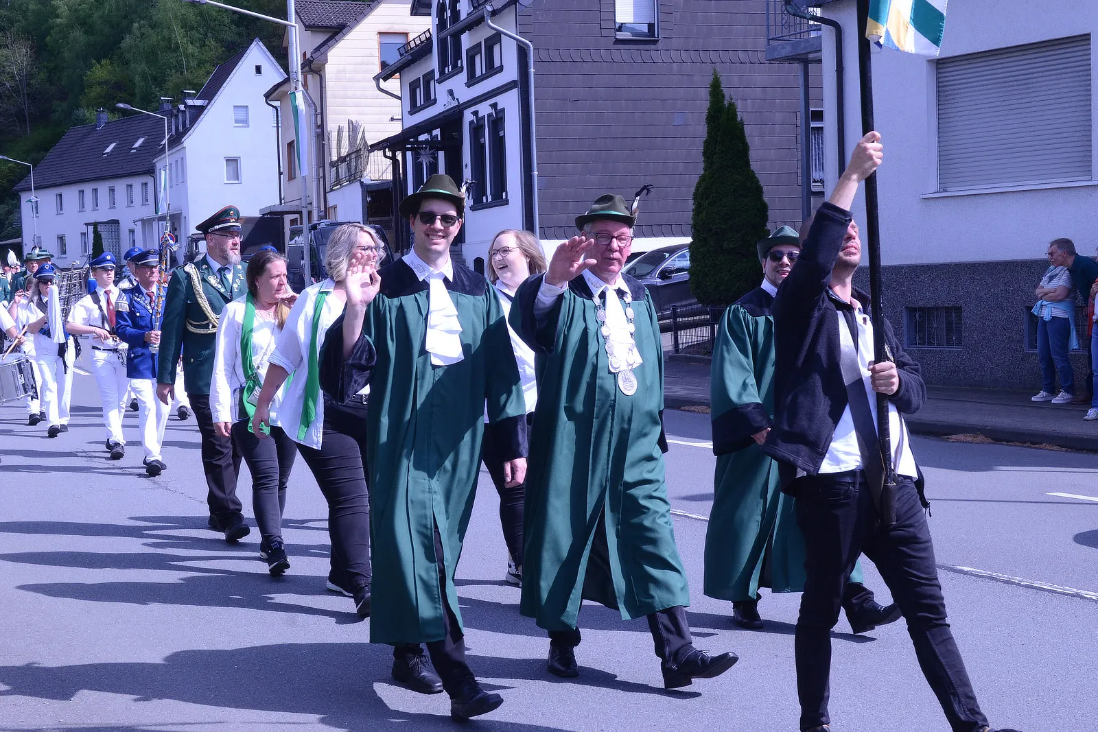 Festumzug mit Musikern in Uniformen und Personen in traditioneller Kleidung, darunter grüne Mäntel und Hüte, zieht an Häusern vorbei.