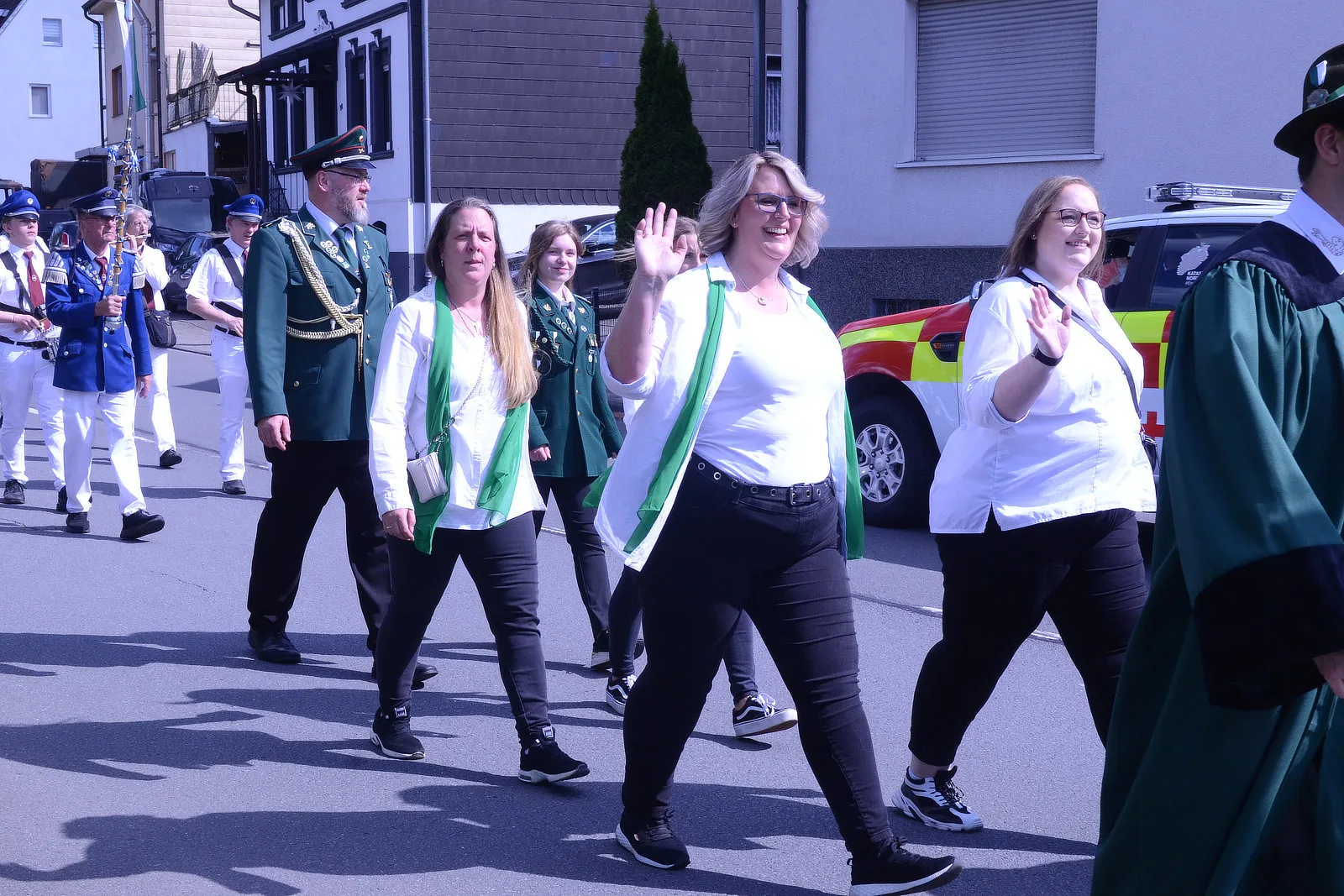 Festumzug mit mehreren Personen in traditioneller Kleidung und grünen Schärpen, die zur Kamera winken. Im Hintergrund eine Musikkapelle und ein rotes Einsatzfahrzeug.