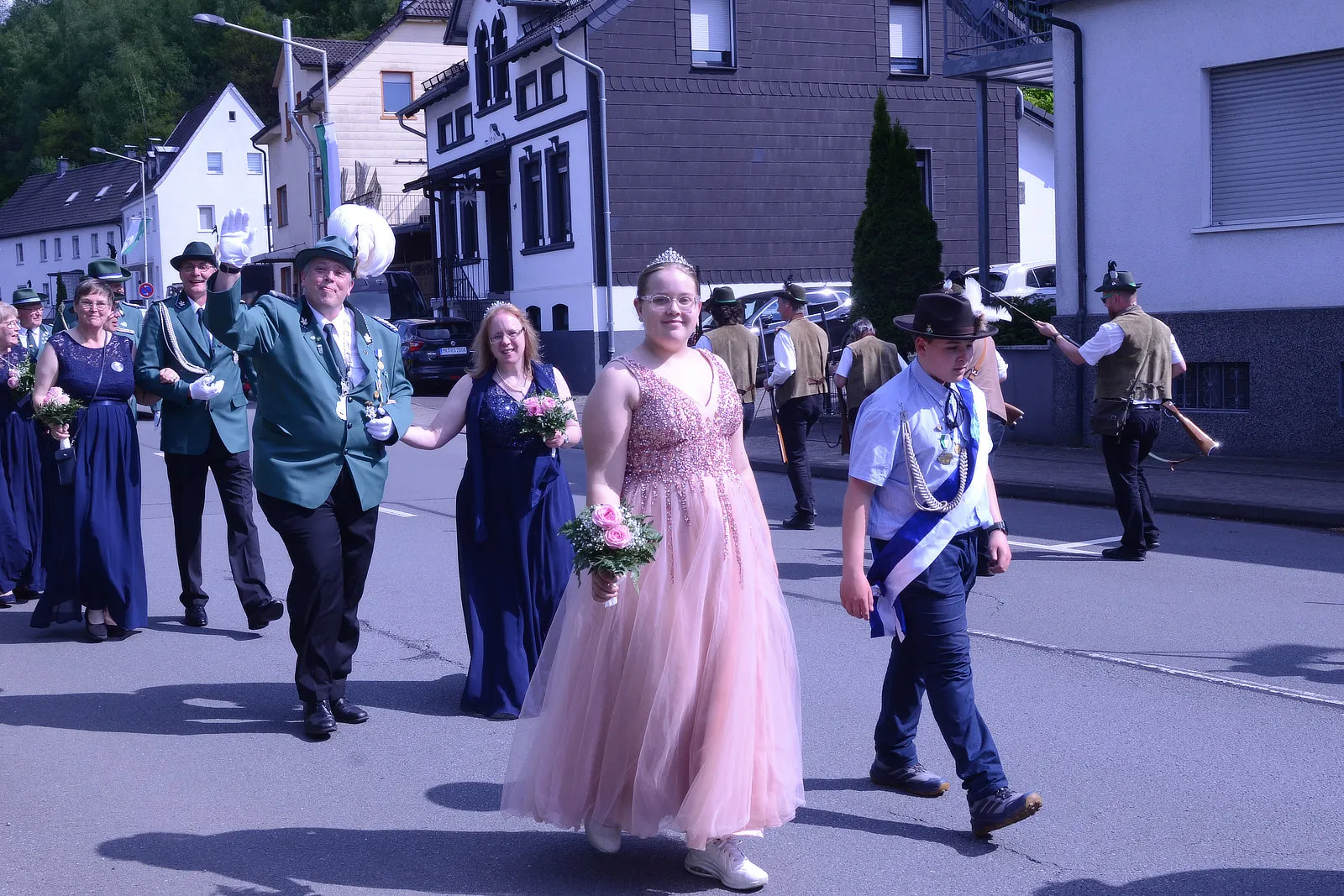 Festumzug mit einer jungen Königin in rosa Ballkleid und Krone, begleitet von Betreuern und einer Musikkapelle.