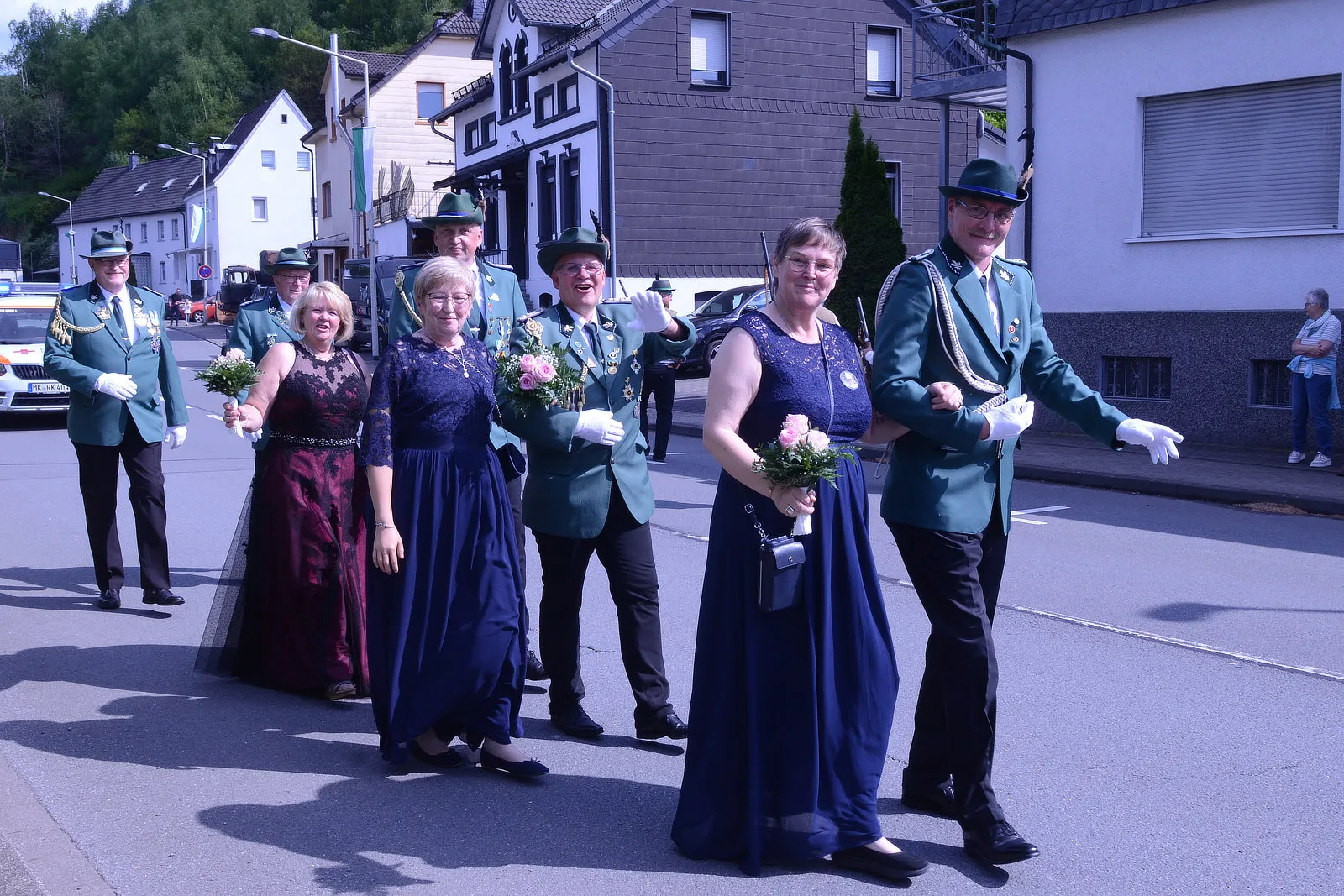 Festumzug mit mehreren Personen in traditioneller Tracht, darunter grüne Uniformen und festliche Kleider, die Blumensträuße tragen und auf einer Straße gehen.
