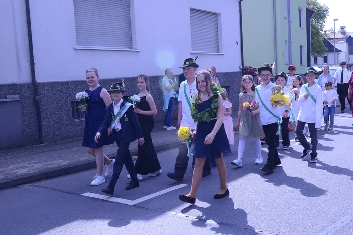 Kinder und Jugendliche ziehen lächelnd in Tracht und mit Blumen durch eine Straße, begleitet von Erwachsenen mit Musikinstrumenten.