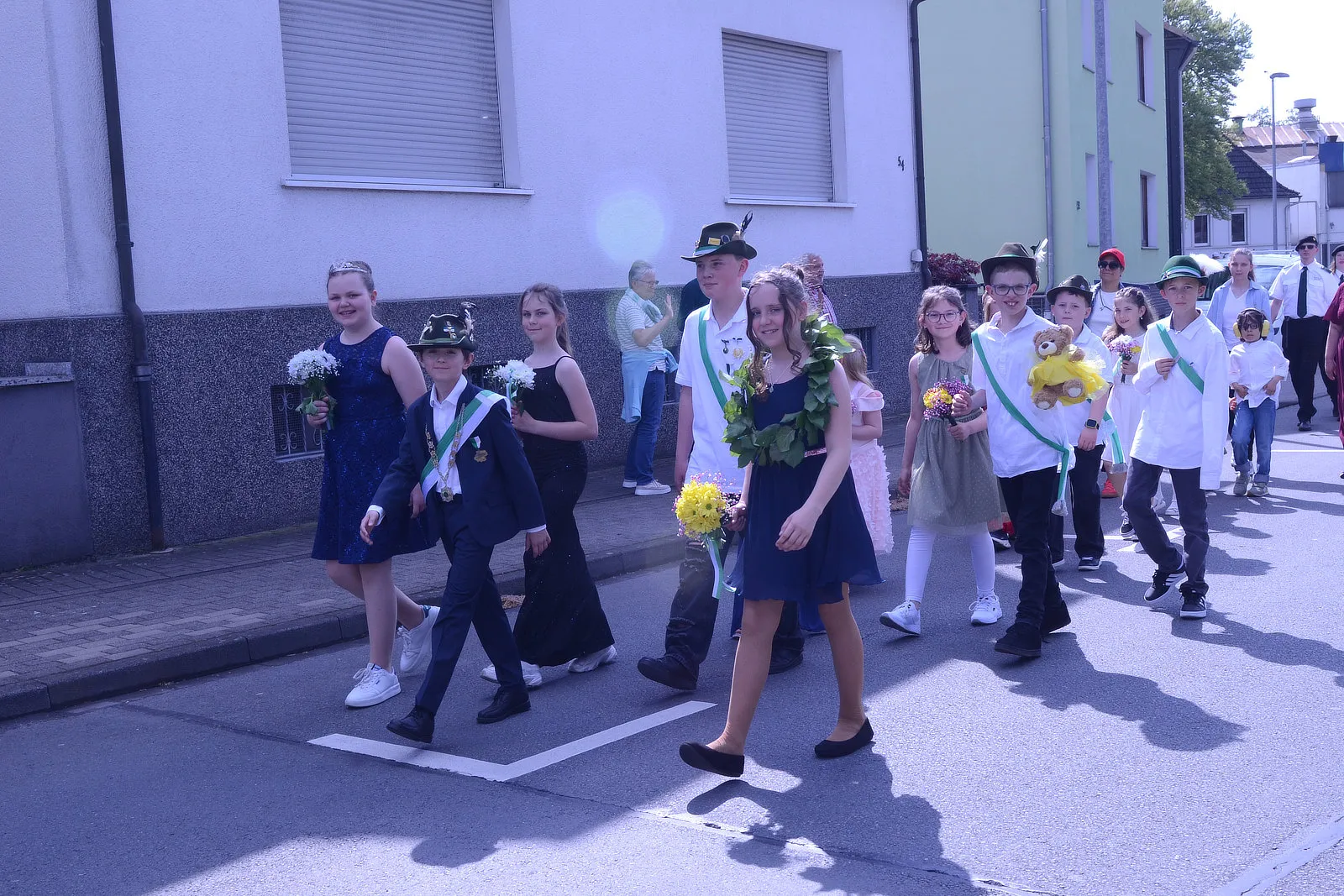 Kinder und Jugendliche ziehen lächelnd in Tracht und mit Blumen durch eine Straße, begleitet von Erwachsenen mit Musikinstrumenten.