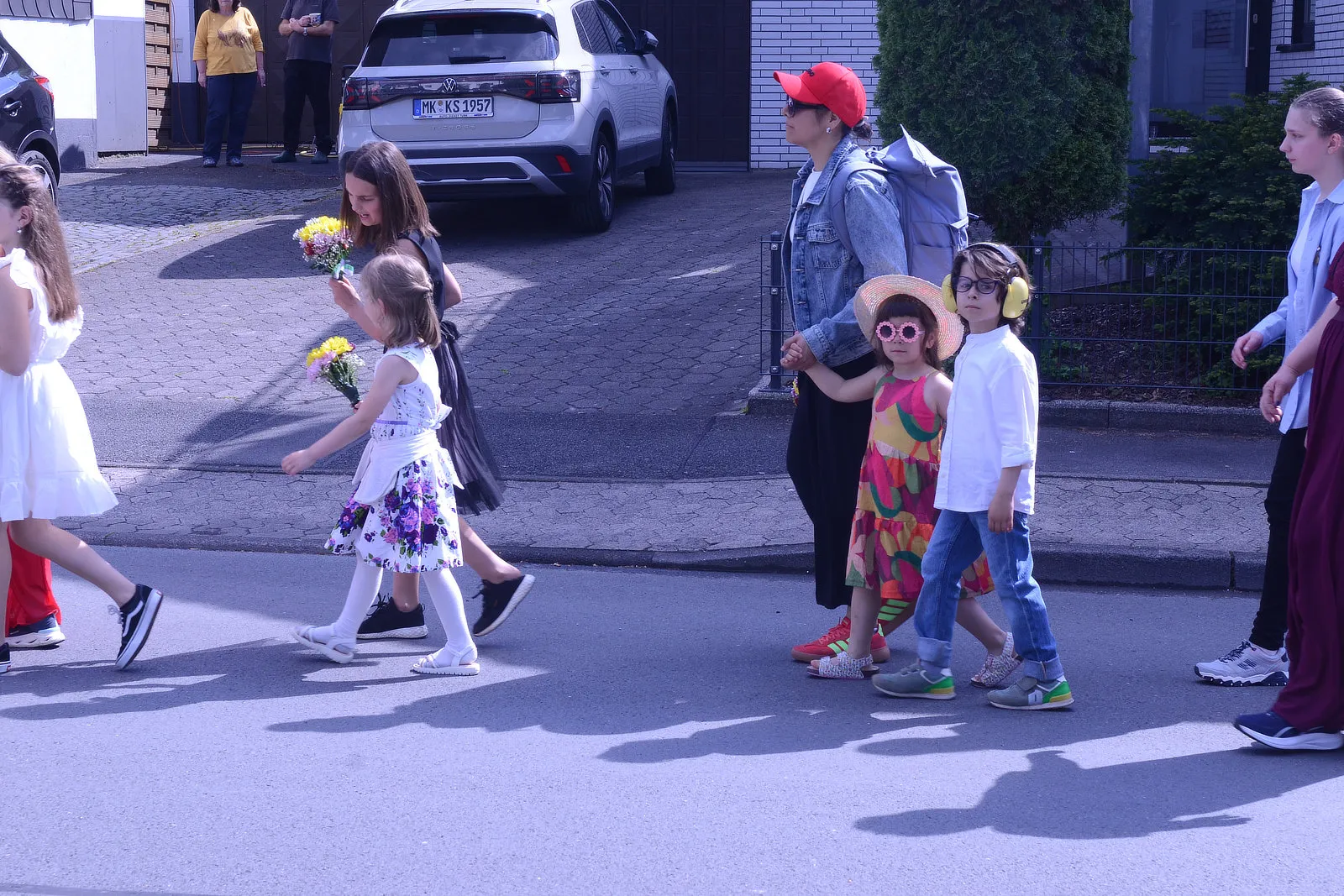 Kinder mit Blumen gehen an einer erwachsenen Person mit roter Kappe und Rucksack über einen Zebrastreifen. Im Hintergrund ein graues Auto und Häuser.