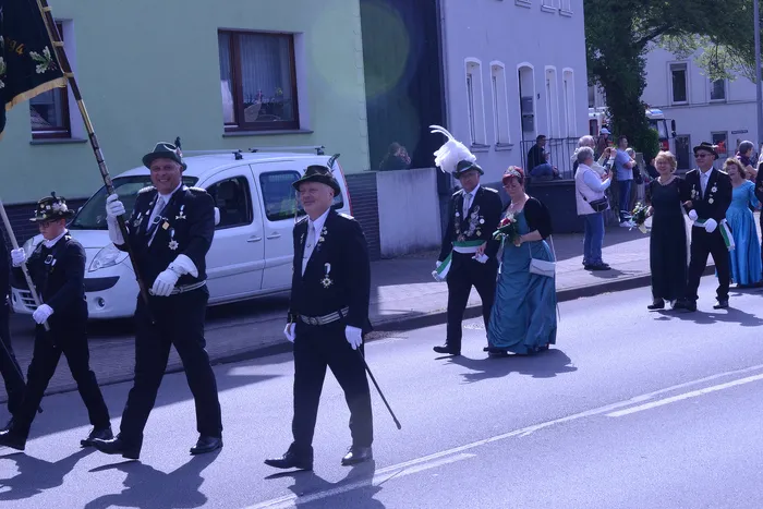 Festumzug mit mehreren Personen in traditioneller Tracht, darunter ein Mann mit Zylinder und eine Frau in einem langen, türkisfarbenen Kleid. Im Hintergrund sind Zuschauer und Gebäude zu sehen.