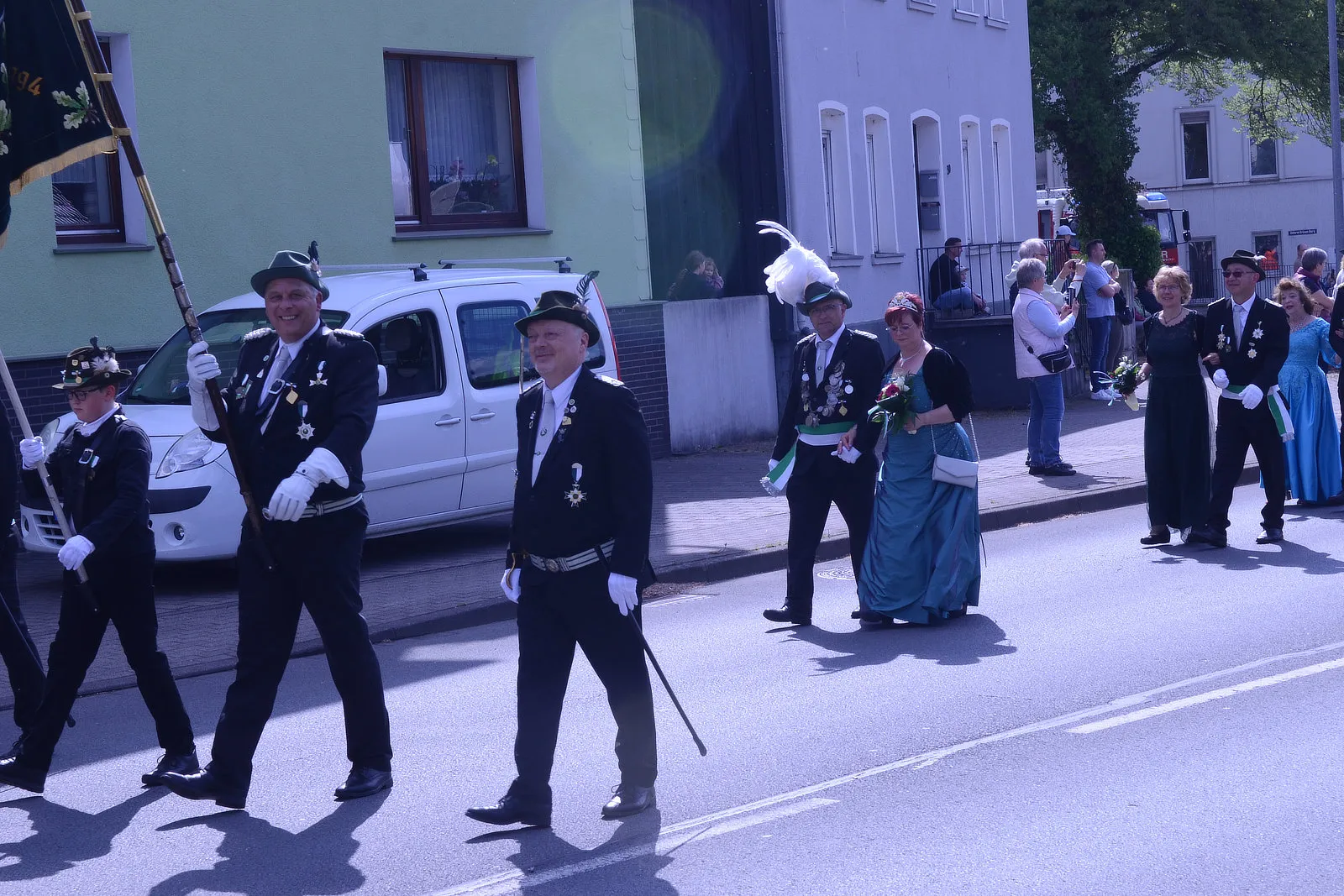 Festumzug mit mehreren Personen in traditioneller Tracht, darunter ein Mann mit Zylinder und eine Frau in einem langen, türkisfarbenen Kleid. Im Hintergrund sind Zuschauer und Gebäude zu sehen.