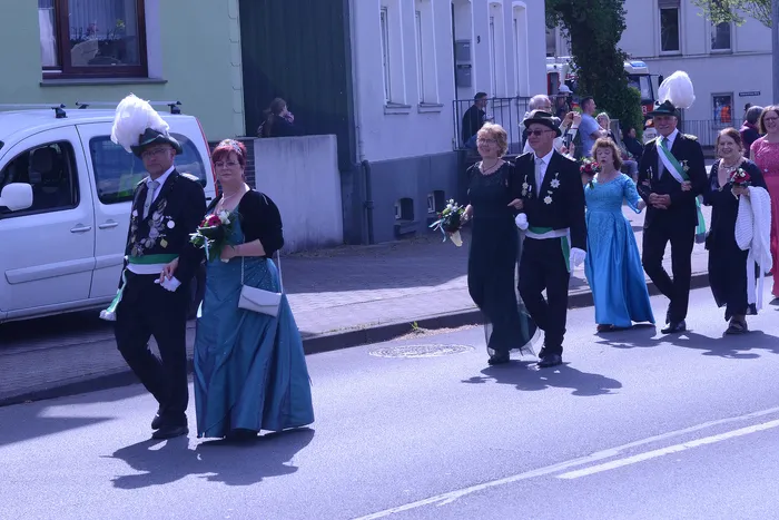 Festumzug mit mehreren Personen in traditioneller Tracht, darunter Uniformen mit hohen Hüten und lange Kleider, die Blumensträuße tragen.