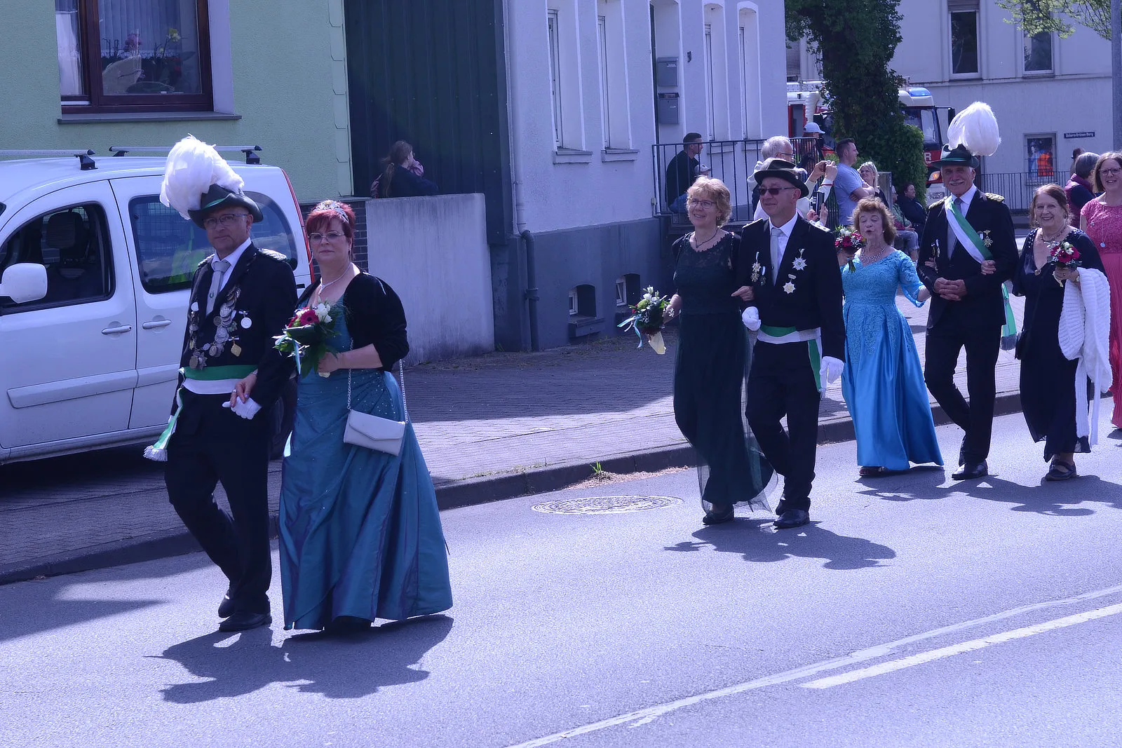 Festumzug mit mehreren Personen in traditioneller Tracht, darunter Uniformen mit hohen Hüten und lange Kleider, die Blumensträuße tragen.