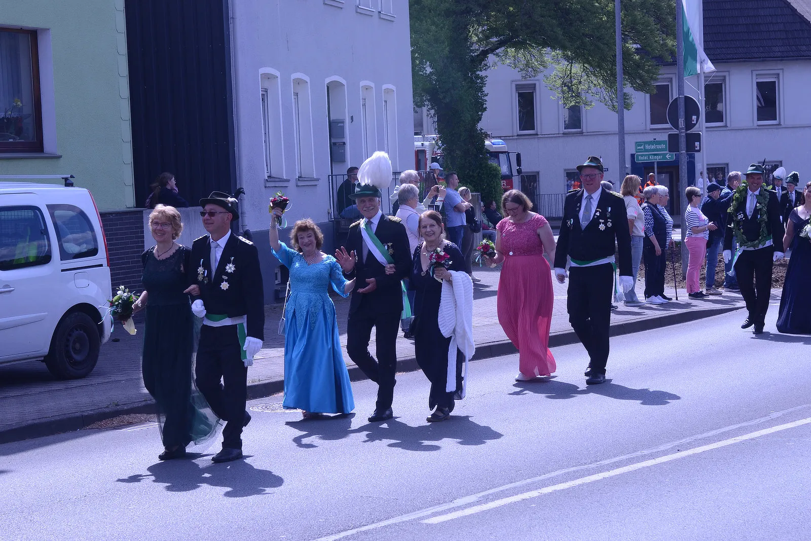 Festumzug mit festlich gekleideten Personen, einige tragen grüne Uniformen und hohe Hüte, andere Abendkleider, und gehen an Zuschauern entlang einer Straße.