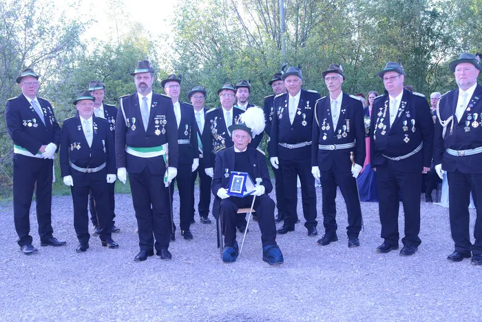 Gruppe von Männern in schwarzer Uniform und Zylinderhüten posiert für ein Foto. Ein Mann sitzt auf einem Stuhl und hält eine Auszeichnung, während die anderen um ihn herum stehen.