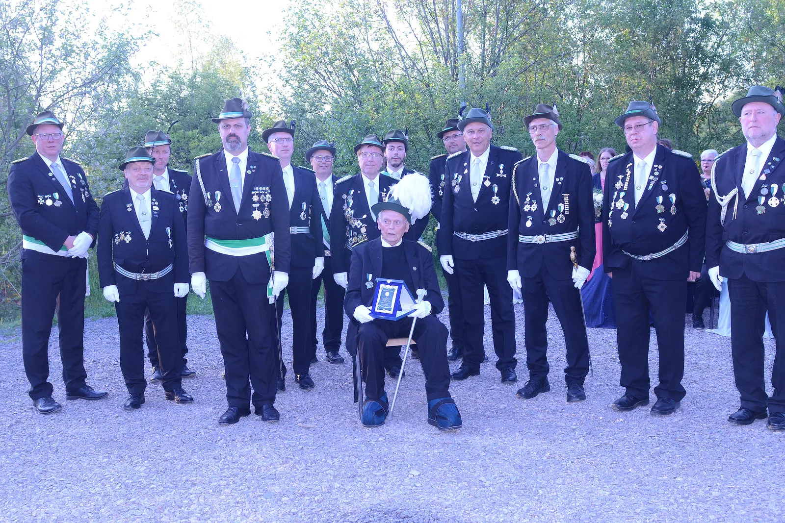 Gruppe von Männern in schwarzer Uniform und Zylinderhüten posiert für ein Foto. Ein Mann sitzt auf einem Stuhl und hält eine Auszeichnung, während die anderen um ihn herum stehen.