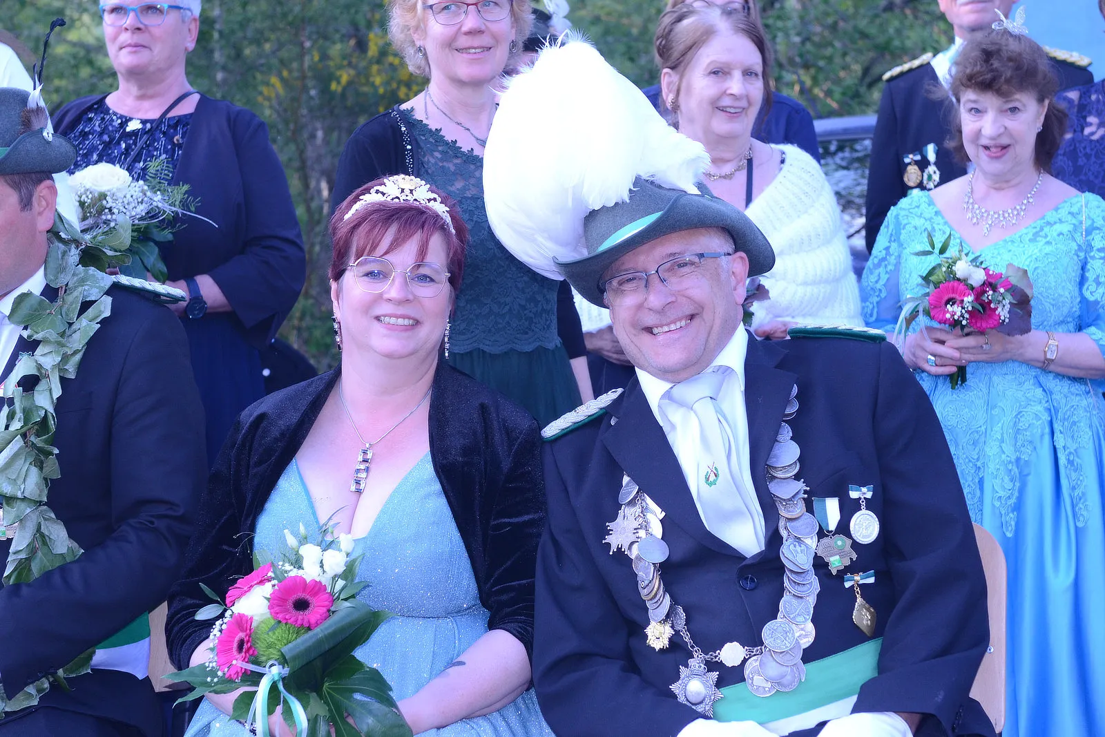 Ein Mann mit Orden und eine Frau in Abendkleidung lächeln in die Kamera, umgeben von weiteren Personen mit Blumen und festlicher Kleidung. Im Hintergrund sind Bäume und Gebäude zu sehen.