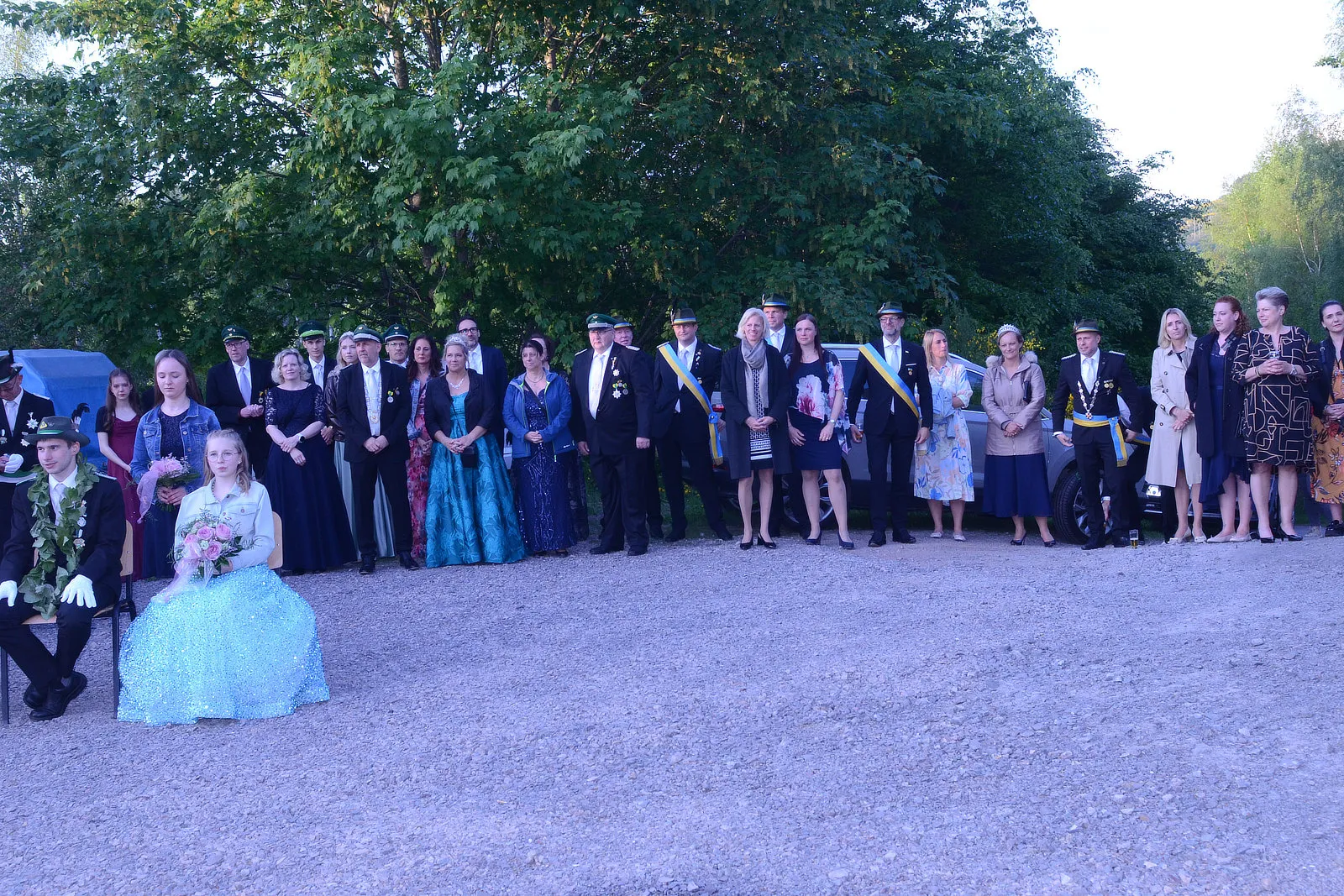 Gruppe von Menschen in festlicher Kleidung steht vor Bäumen, darunter ein Mädchen in einem blauen Kleid und ein Mann in Uniform mit einem Blumenkranz.