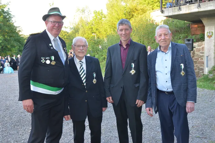 Vier Männer in festlicher Kleidung mit Orden posieren im Freien vor einer Mauer und Bäumen. Im Hintergrund sind weitere Menschen zu sehen, die zu einem Schützenfest gehören.