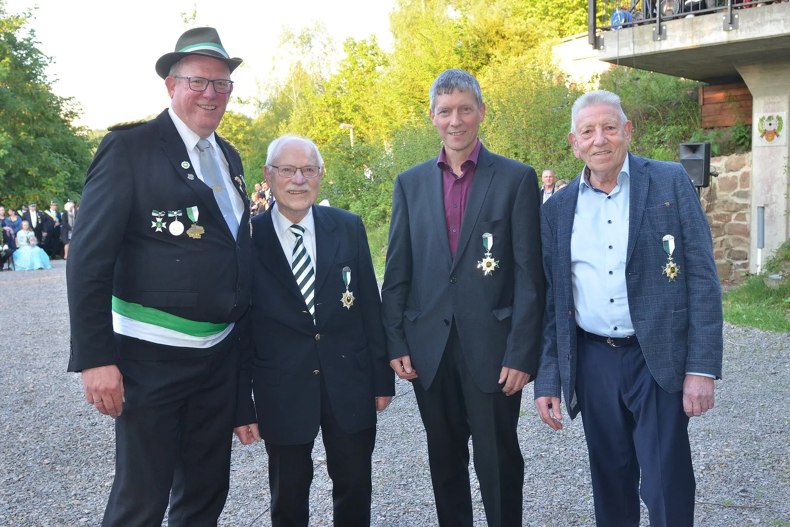 Vier Männer in festlicher Kleidung mit Orden posieren im Freien vor einer Mauer und Bäumen. Im Hintergrund sind weitere Menschen zu sehen, die zu einem Schützenfest gehören.
