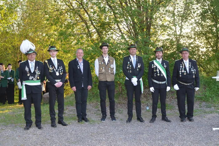 Sieben Männer in festlicher Tracht stehen in einer Reihe vor Bäumen. Sie tragen grüne Uniformen mit goldenen Verzierungen und Medaillen, einige auch grüne Hüte. Im Hintergrund sind weitere Personen in Uniformen zu sehen.