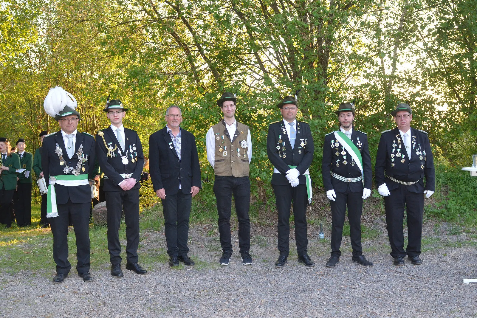 Sieben Männer in festlicher Tracht stehen in einer Reihe vor Bäumen. Sie tragen grüne Uniformen mit goldenen Verzierungen und Medaillen, einige auch grüne Hüte. Im Hintergrund sind weitere Personen in Uniformen zu sehen.
