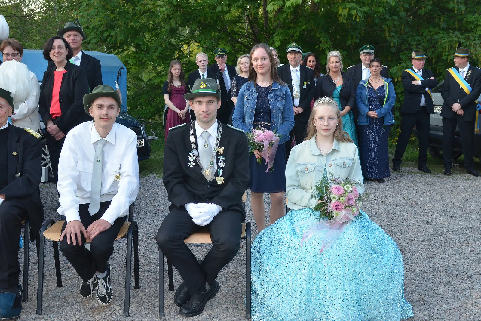 Drei junge Erwachsene im festlichen Gewand stehen im Mittelpunkt einer Gruppe von Menschen in Uniformen und zivilen Kleidern. Im Hintergrund ein Pferd und Bäume.