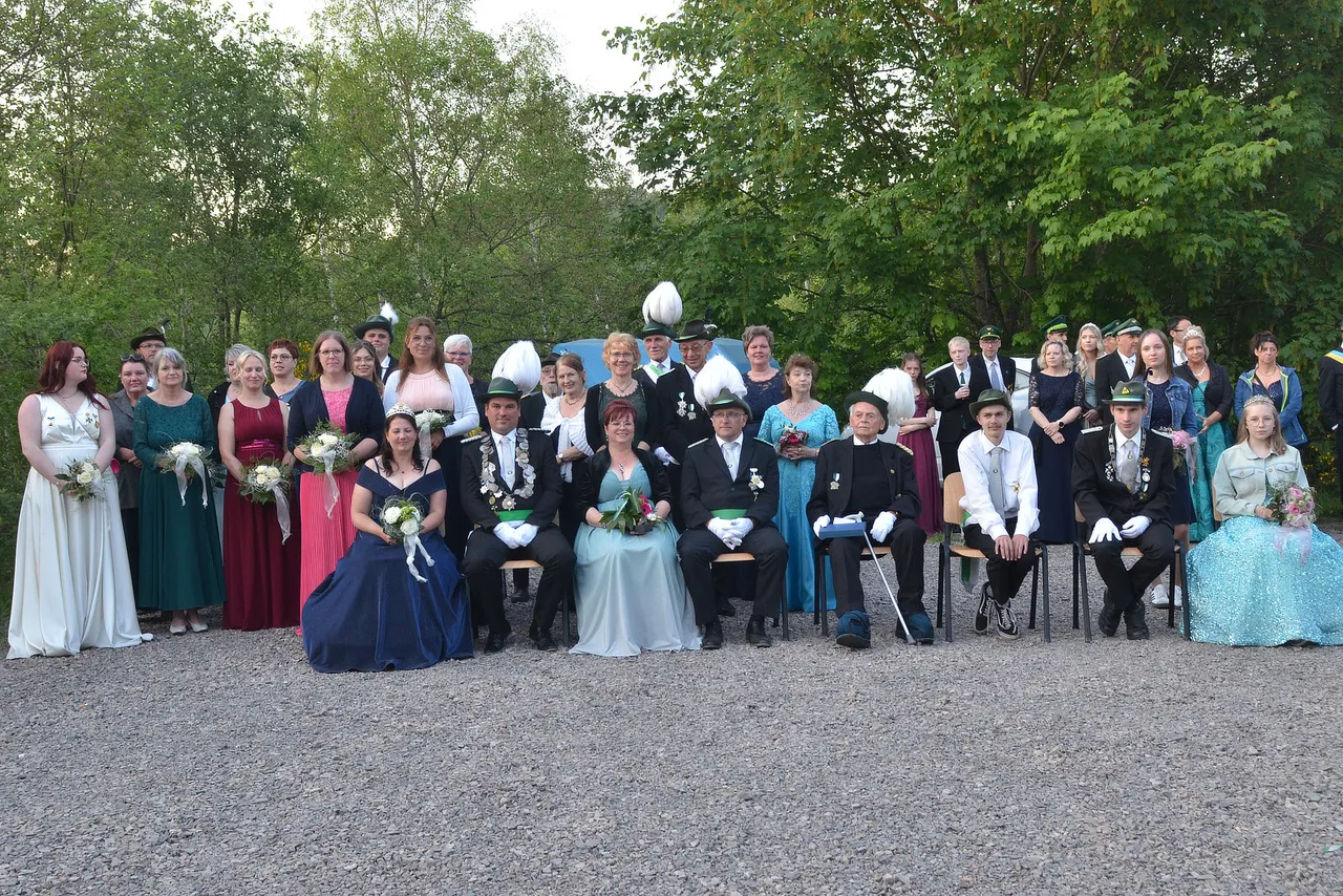 Gruppe von etwa 30 Personen in festlicher Kleidung posiert für ein Gruppenfoto vor Bäumen. Einige tragen Schützenuniformen mit hohen Hüten, andere Abendgarderobe.