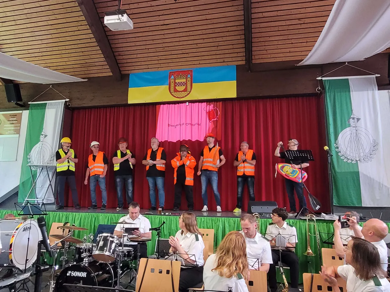 Chor mit Warnwesten singt auf einer Bühne vor rotem Vorhang mit Wappen, darunter Musiker mit Blasinstrumenten und Schlagzeug.