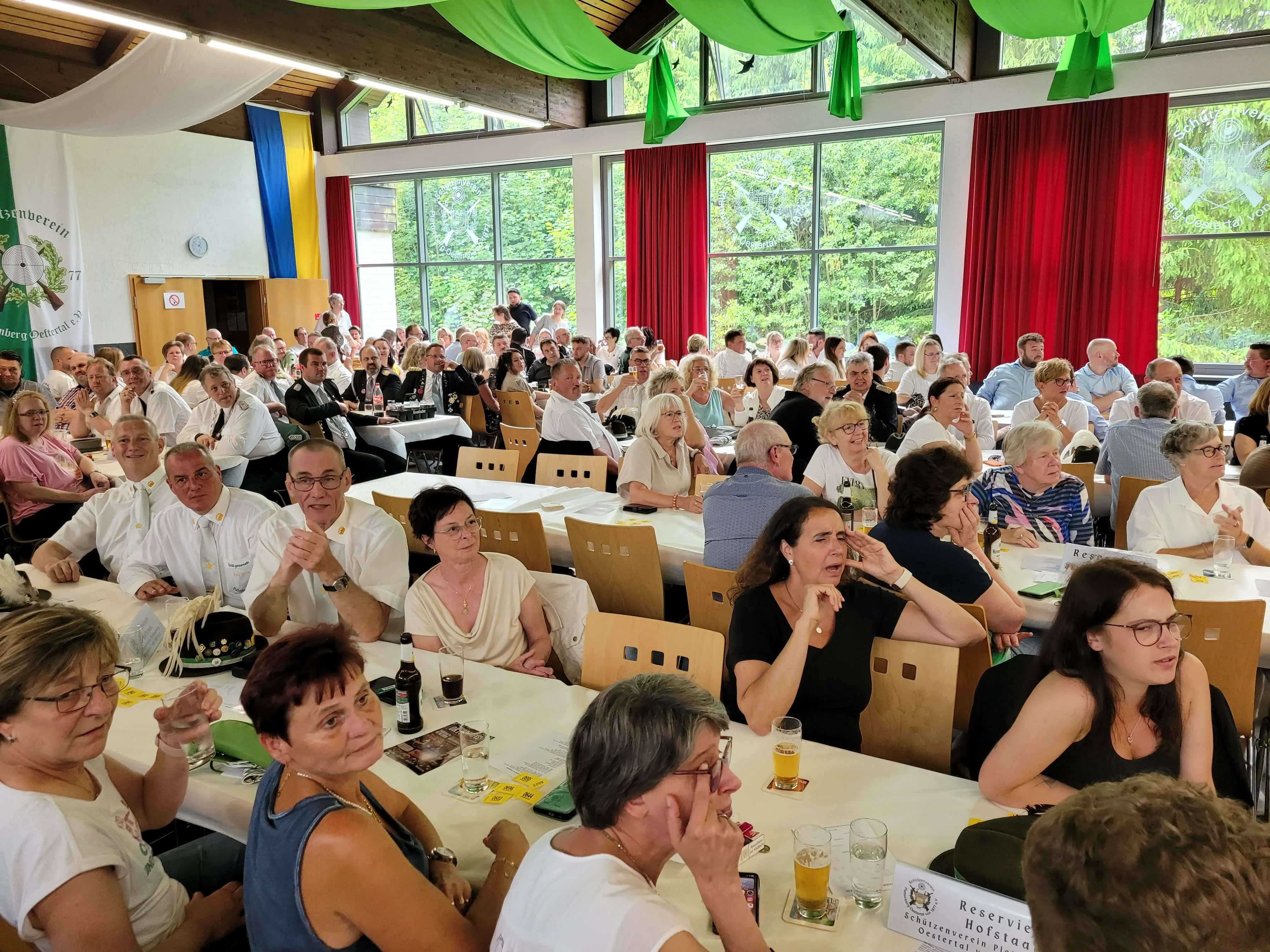 Große Menschenmenge bei einem festlichen Anlass in einem hellen Raum mit Holztäfelung und großen Fenstern. Einige Personen sitzen an Tischen mit Getränken, andere stehen und unterhalten sich.