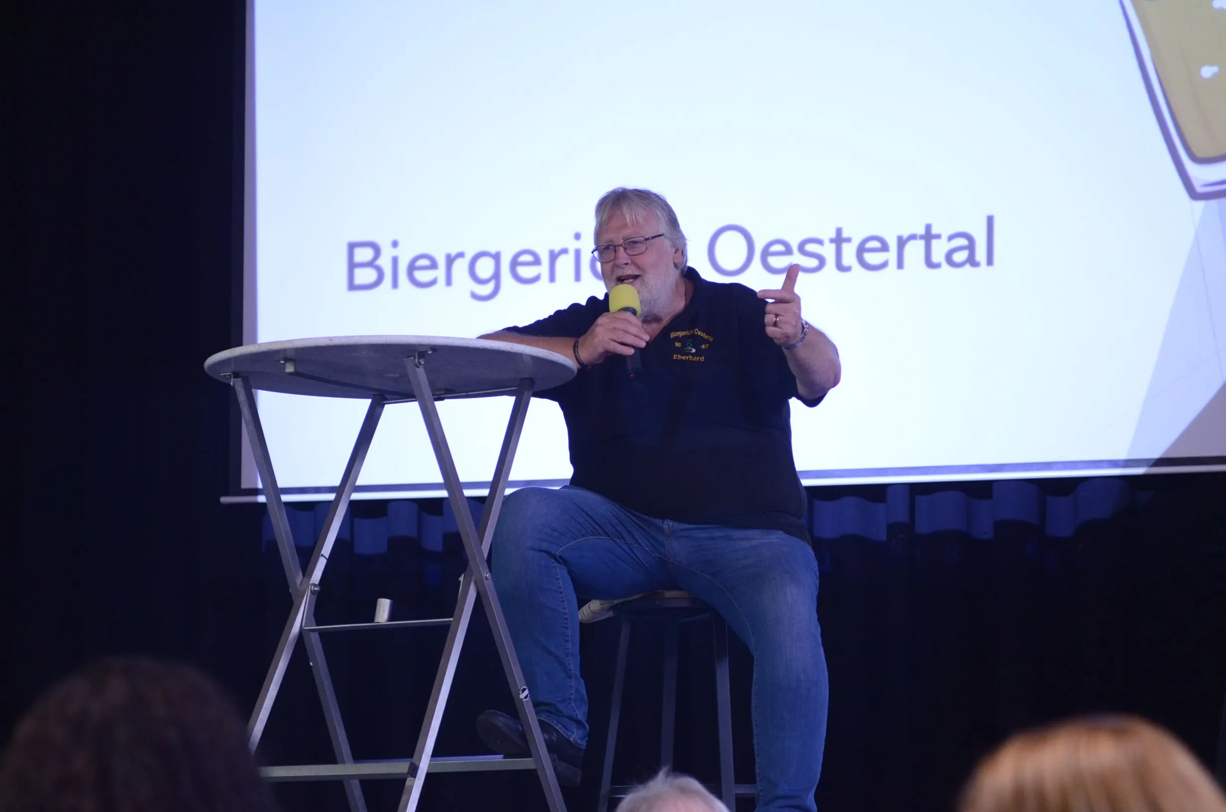 Ein Mann mit Bart hält ein Mikrofon in der Hand und spricht auf einer Bühne. Im Hintergrund eine Projektion mit der Aufschrift „Biergerie Oestertal“.