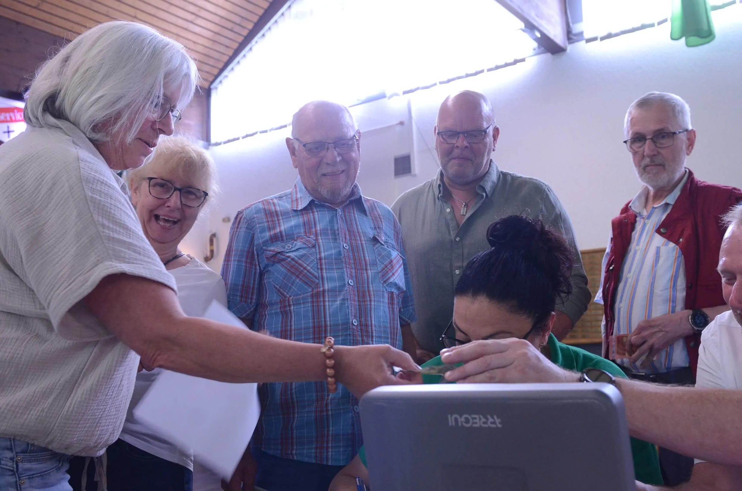 Mehrere Menschen stehen um einen Laptop versammelt, eine Frau zeigt auf den Bildschirm. Im Hintergrund ein Raum mit Holzbalken und ein rotes Kreuz.