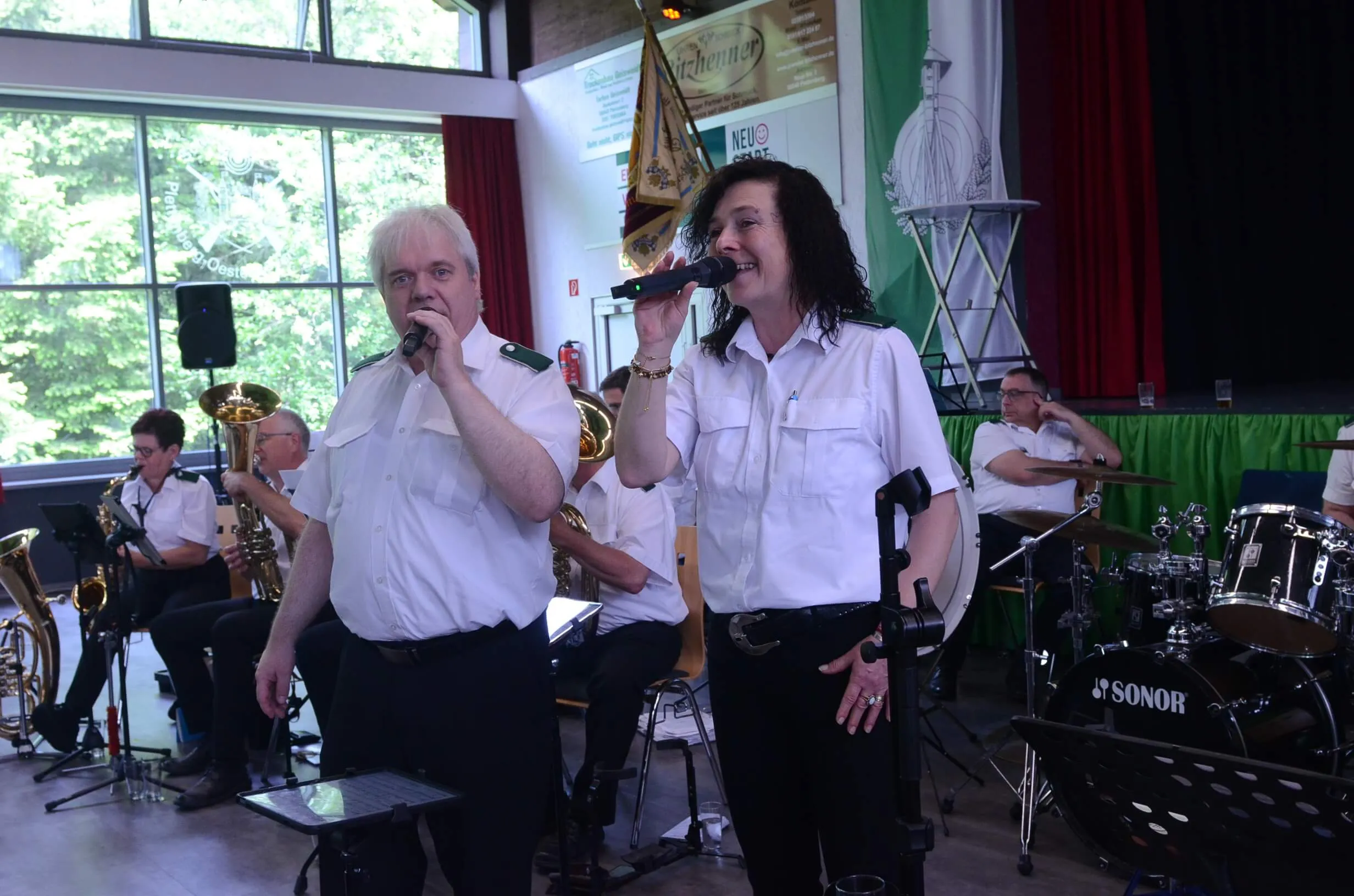 Zwei Personen singen in Mikrophone auf einer Bühne, begleitet von einer Blaskapelle. Im Hintergrund eine Fensterfront und Banner.
