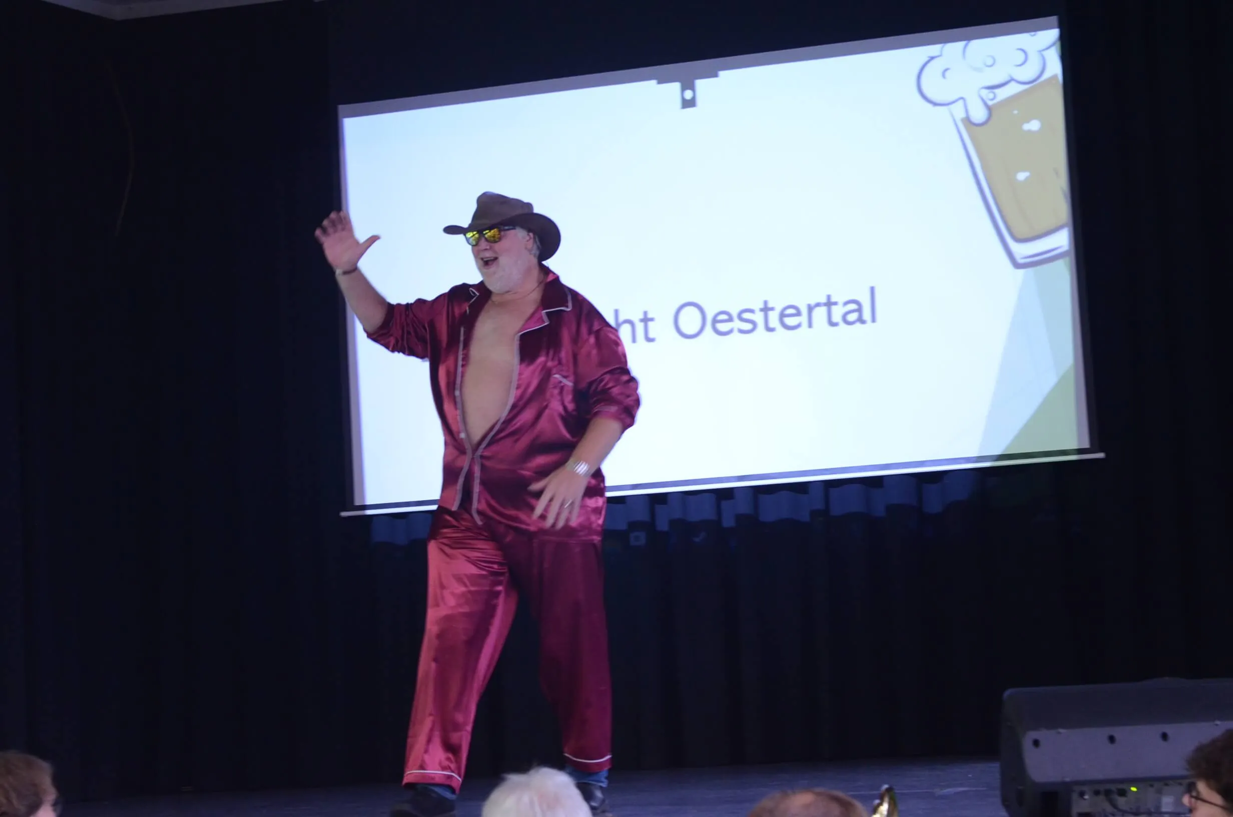 Mann in rotem Anzug und Cowboyhut steht auf einer Bühne und winkt, während im Hintergrund eine Präsentation mit dem Text „Oestertal“ und einem Bierglas zu sehen ist.
