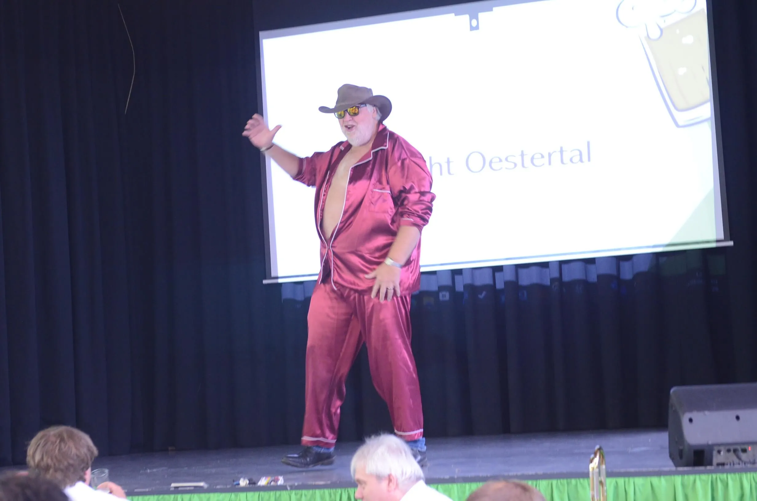 Ein Mann in rotem Pyjama und Cowboyhut steht auf einer Bühne und gestikuliert mit einer Hand. Im Hintergrund eine Projektion mit dem Schriftzug „ht Oestertal“.