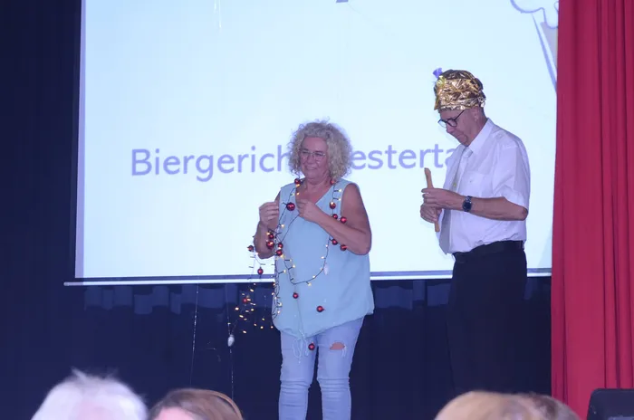 Zwei Personen auf einer Bühne, eine Frau mit einer Lichterkette und ein Mann mit goldener Kopfbedeckung, vor einer Projektion mit dem Text „Biergericht“.