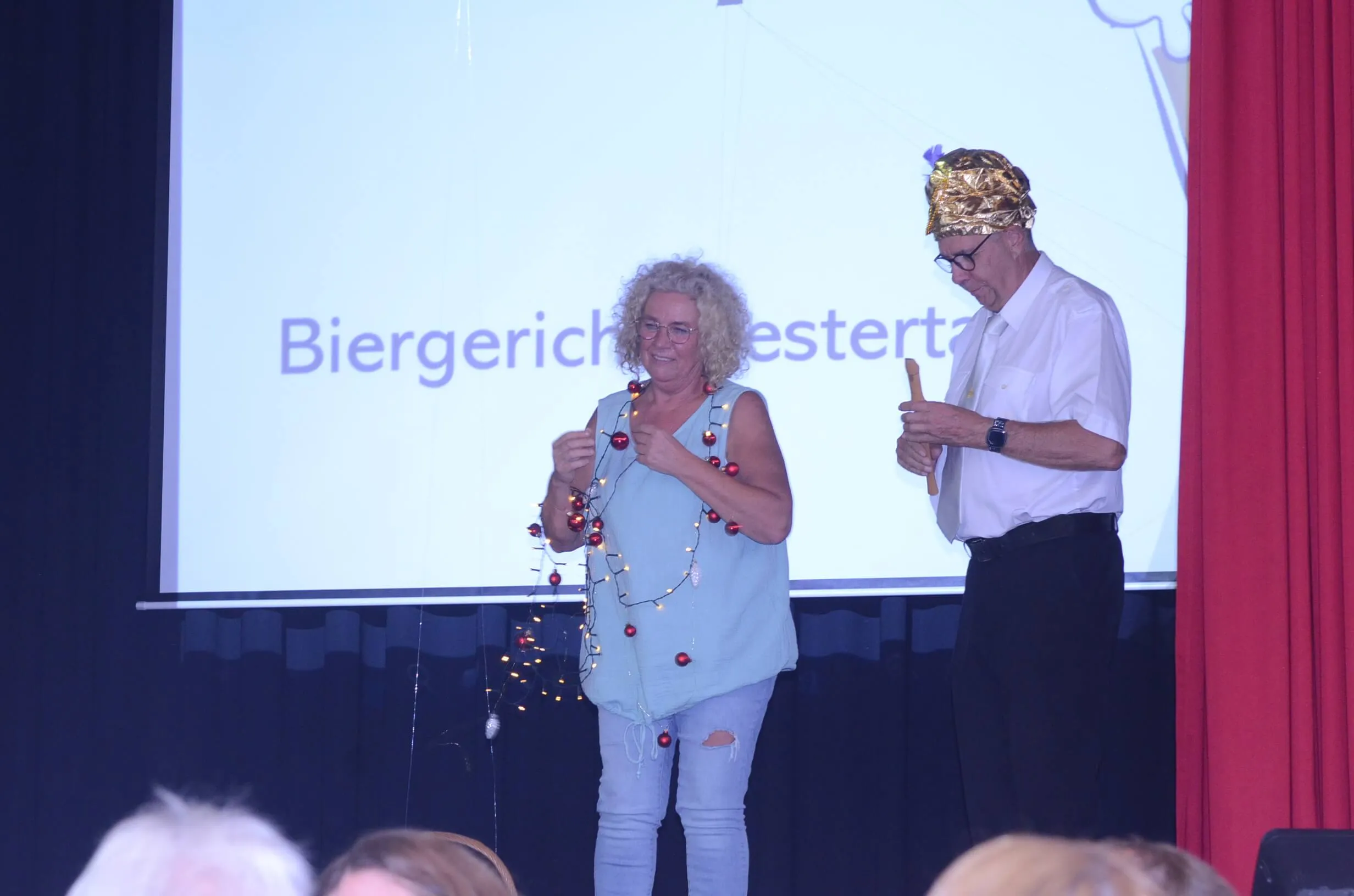 Zwei Personen auf einer Bühne, eine Frau mit einer Lichterkette und ein Mann mit goldener Kopfbedeckung, vor einer Projektion mit dem Text „Biergericht“.