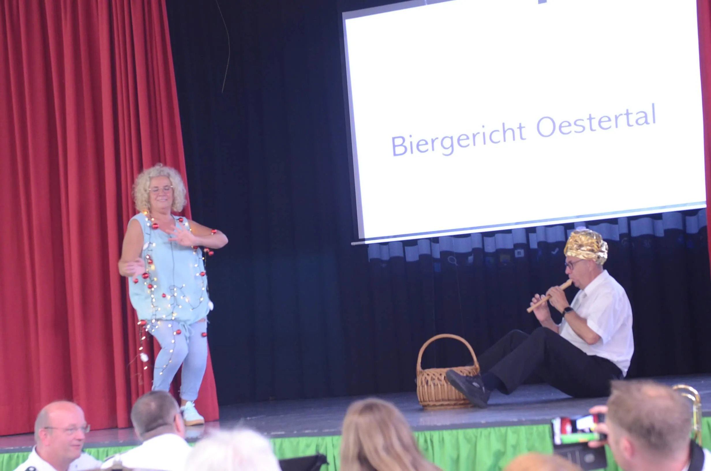 Zwei Personen in festlicher Kleidung spielen auf einer Bühne vor rotem Vorhang und Publikum. Im Hintergrund eine Leinwand mit der Aufschrift „Biergericht Oestertal“.