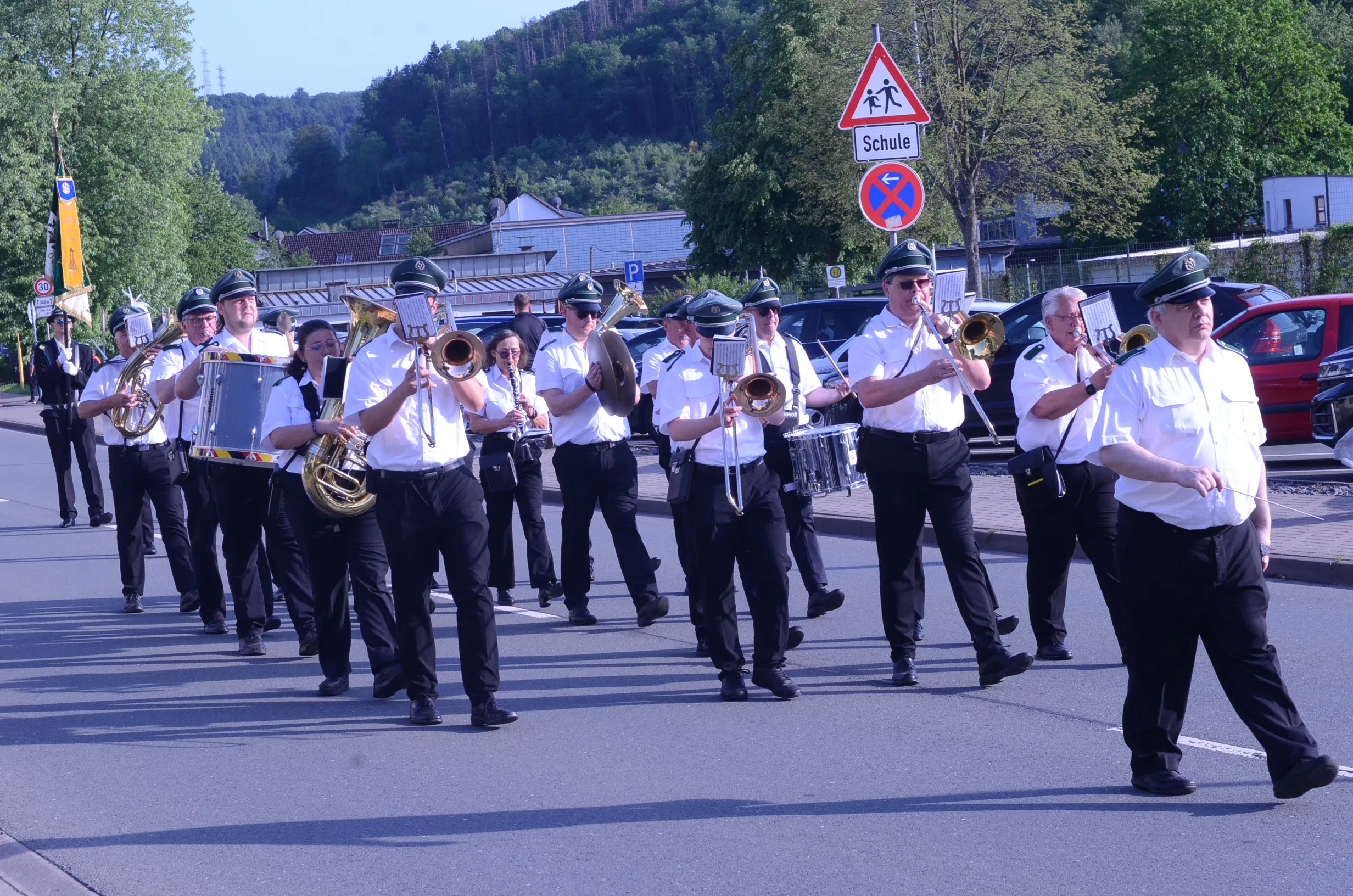 Musikzug mit Trompeten, Posaunen, Klarinetten und Trommeln marschiert auf einer Straße, im Hintergrund ein Schild mit der Aufschrift „Schule“.