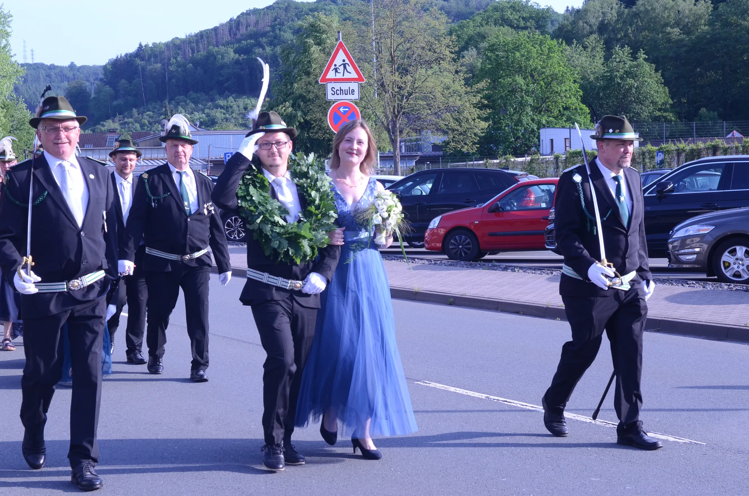 Festzug in Oestertal mit einer Königin in blauem Kleid, die von zwei Männern in Uniformen mit Hüten begleitet wird. Im Hintergrund sind weitere Personen in Uniformen und geparkte Autos zu sehen.