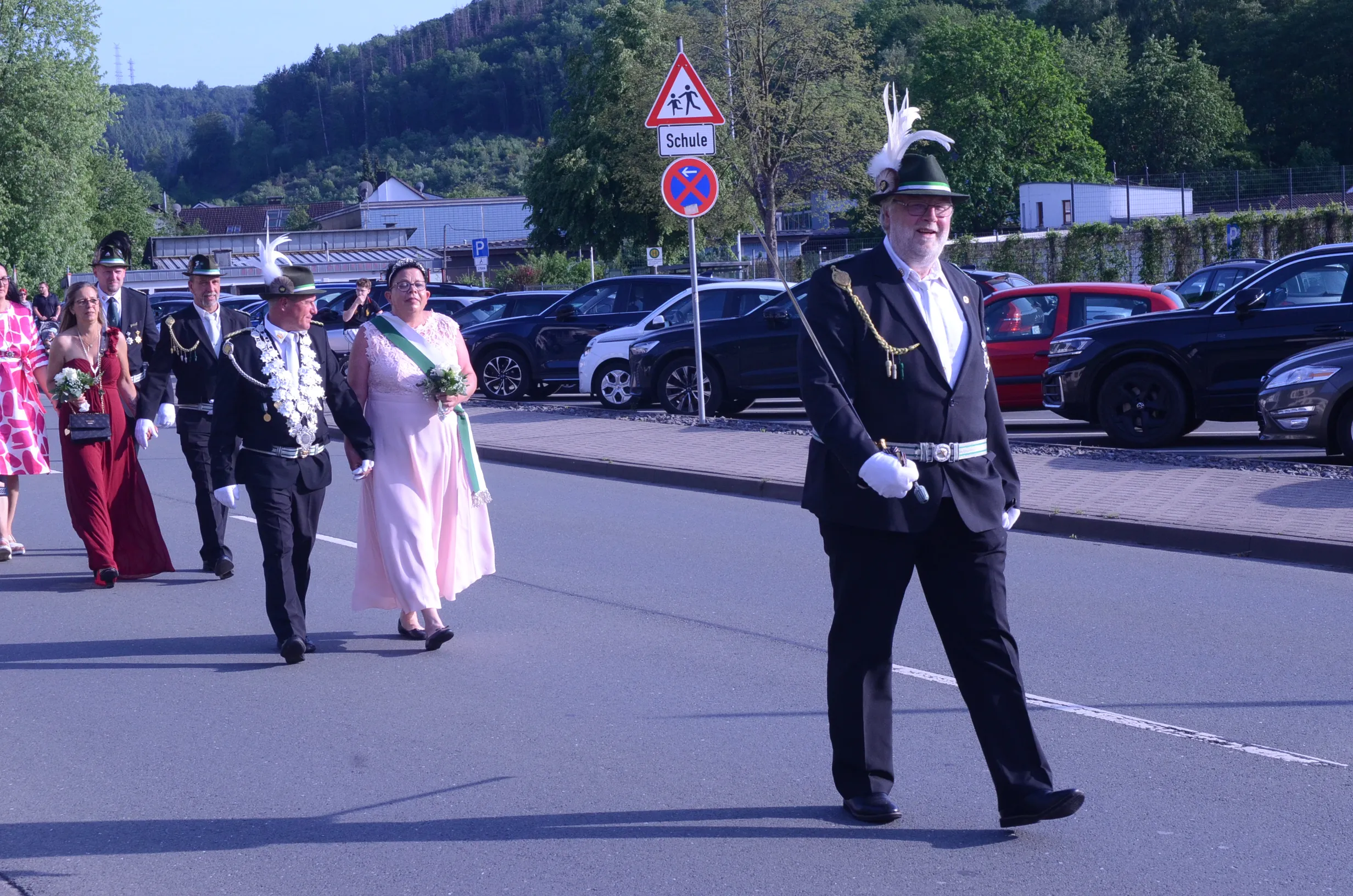 Festzug in Oestertal: Ein Mann in Uniform und Zylinder führt eine Gruppe von Menschen in traditioneller Tracht an, während sie auf einer Straße marschieren. Im Hintergrund sind geparkte Autos und ein Straßenschild zu sehen.