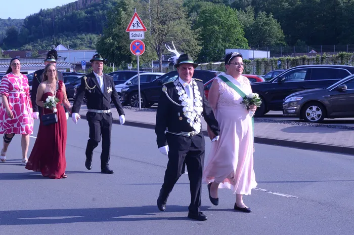 Festzug in Oestertal mit festlich gekleideten Personen, darunter ein Mann in Uniform und eine Frau mit Blumenstrauß, vor einer Straße mit parkenden Autos und einem Schulwegweiser.
