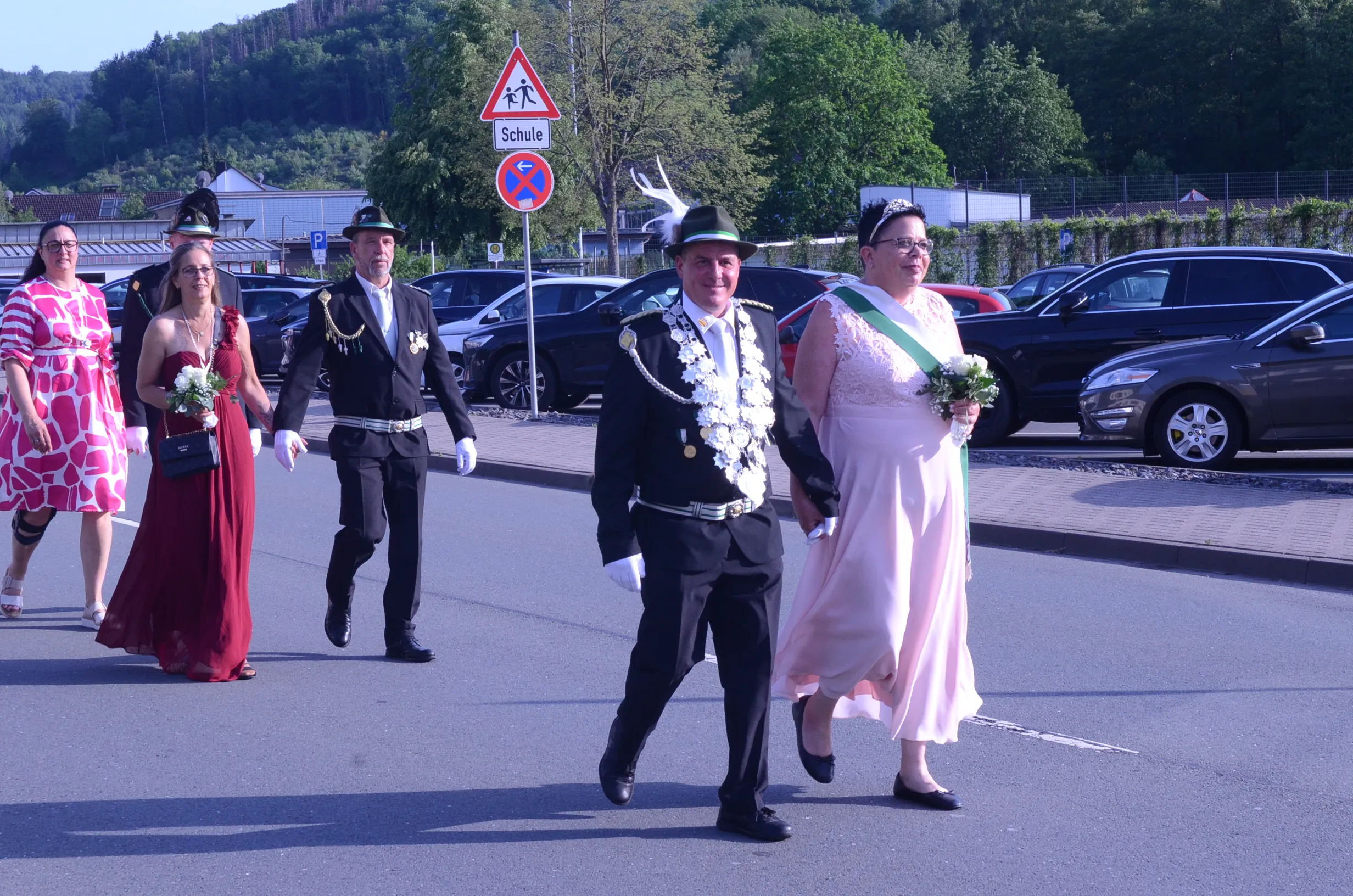 Festzug in Oestertal mit festlich gekleideten Personen, darunter ein Mann in Uniform und eine Frau mit Blumenstrauß, vor einer Straße mit parkenden Autos und einem Schulwegweiser.