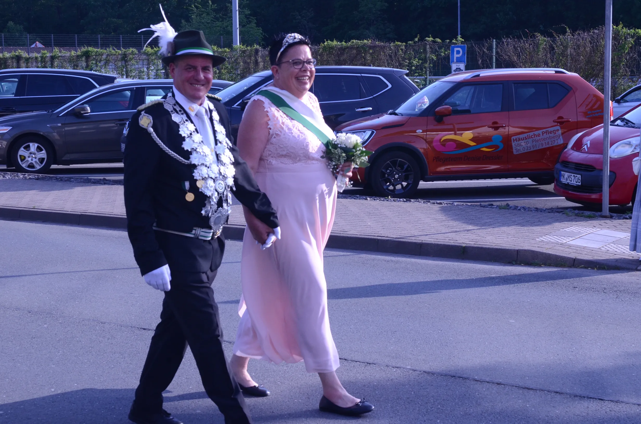Ein Mann in festlicher Uniform und eine Frau in einem hellrosa Kleid gehen lächelnd nebeneinander, die Frau hält einen Blumenstrauß. Im Hintergrund sind geparkte Autos und ein Transporter mit der Aufschrift „Häusliche Pflege“ zu sehen.