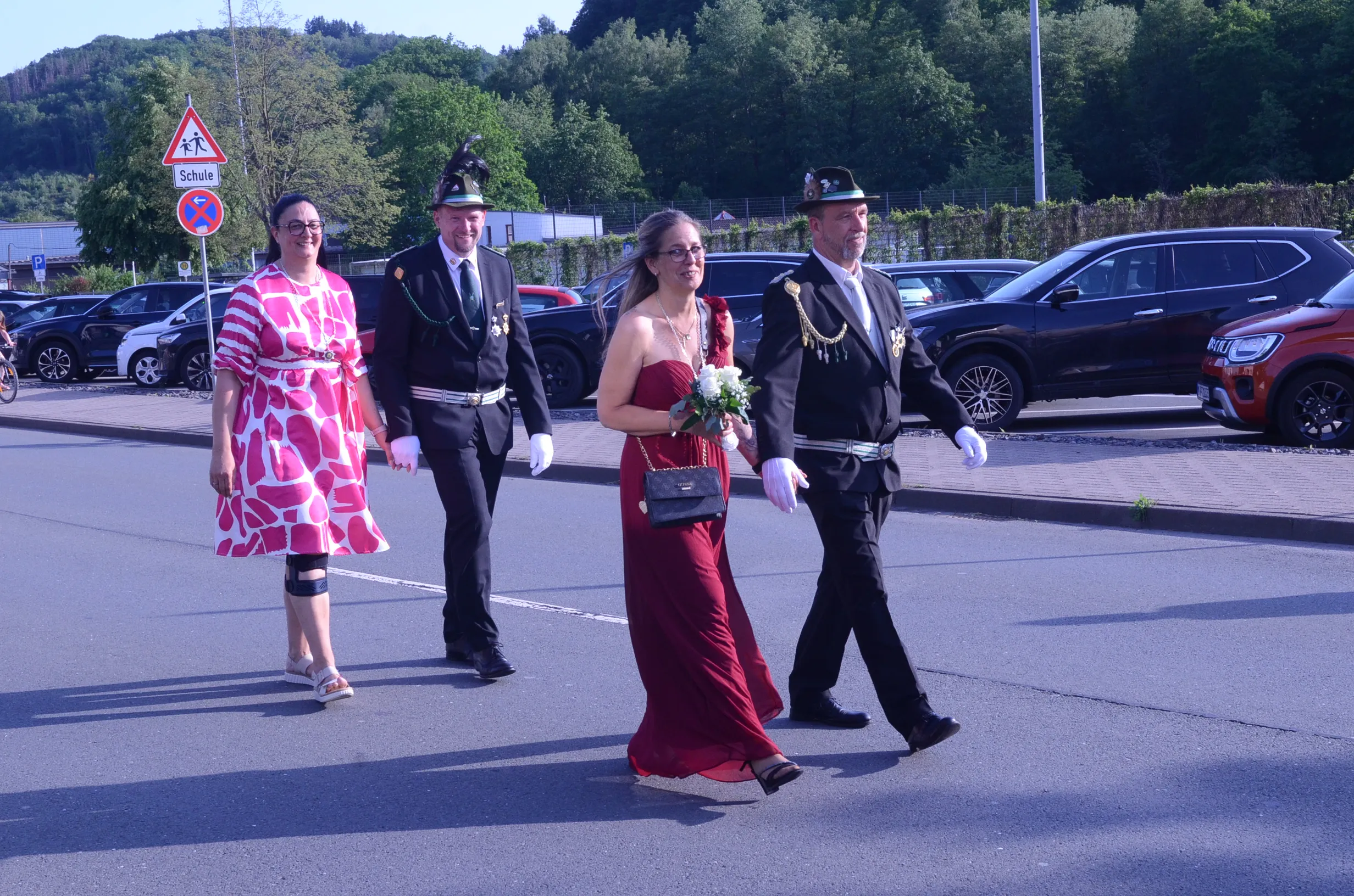 Zwei festlich gekleidete Paare schreiten während eines Schützenfestumzugs über eine Straße. Die Frau trägt ein rotes Abendkleid, der Mann eine Uniform mit weißer Handschuhen und einen hohen Hut.