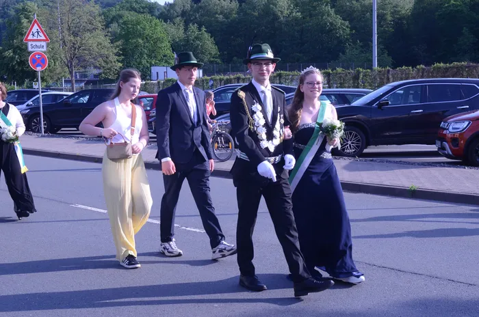 Vier junge Menschen schreiten auf einer Straße, zwei von ihnen tragen festliche Kränze und eine Krone, während die anderen formelle Kleidung tragen. Im Hintergrund sind geparkte Autos und Bäume zu sehen.
