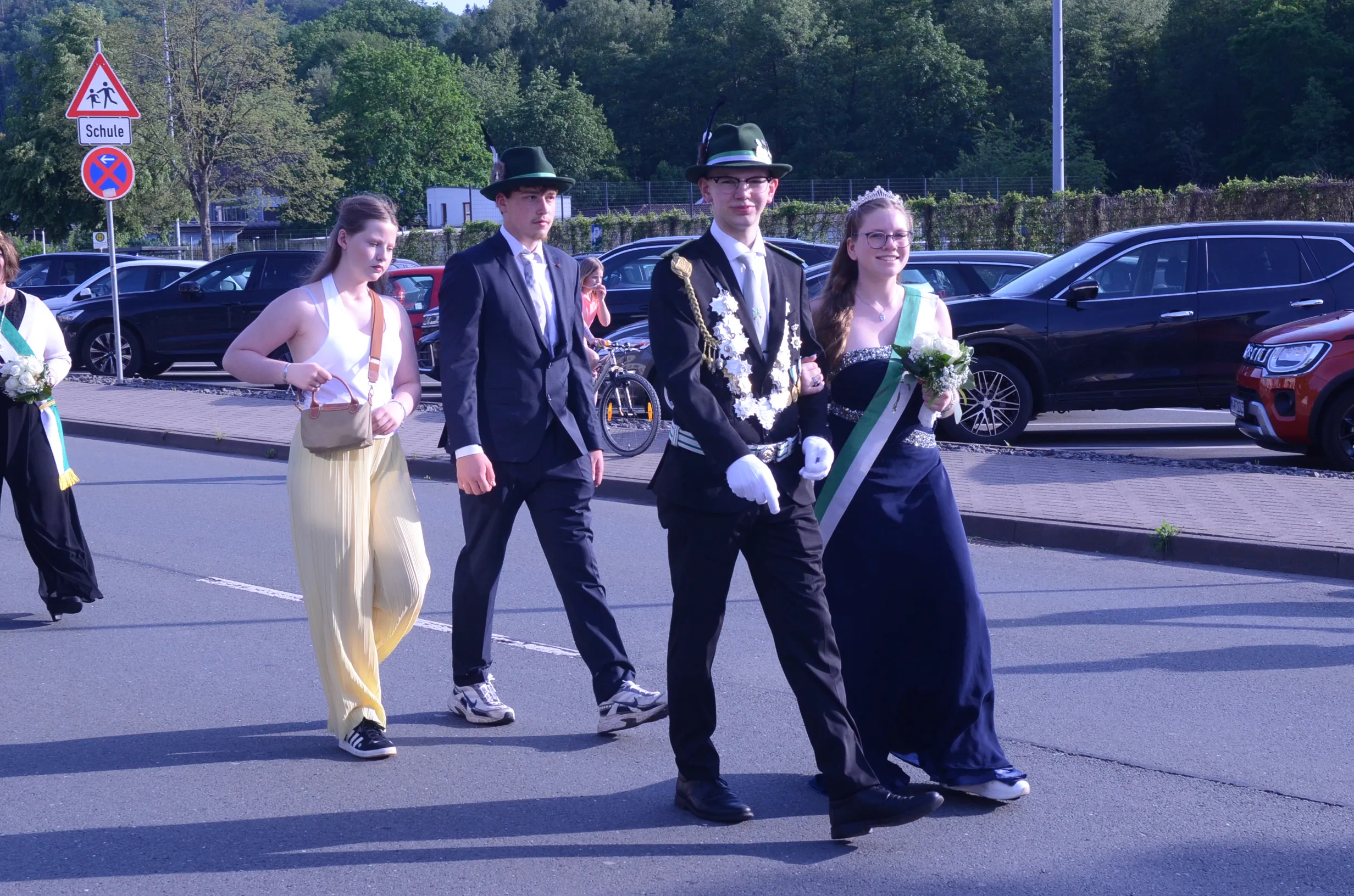 Vier junge Menschen schreiten auf einer Straße, zwei von ihnen tragen festliche Kränze und eine Krone, während die anderen formelle Kleidung tragen. Im Hintergrund sind geparkte Autos und Bäume zu sehen.
