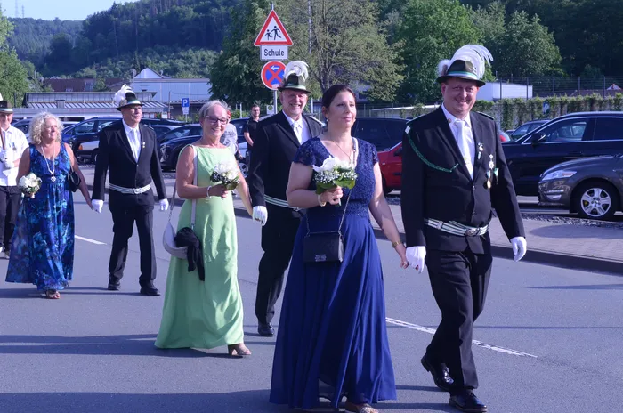 Festzugsteilnehmer in traditioneller Tracht gehen auf einer Straße, einige tragen Blumensträuße. Im Hintergrund ein Verkehrsschild mit dem Hinweis auf eine Schule.