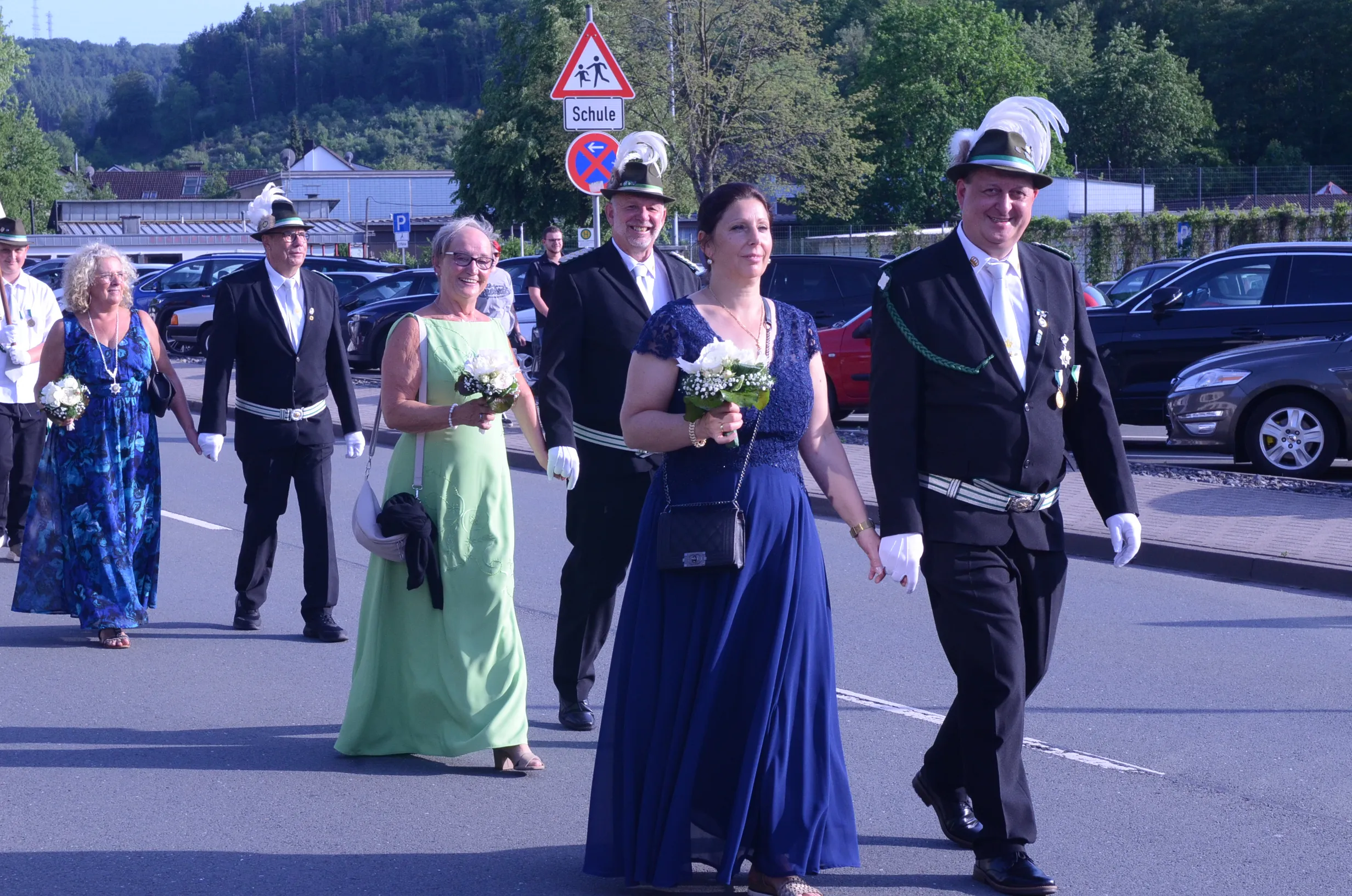 Festzugsteilnehmer in traditioneller Tracht gehen auf einer Straße, einige tragen Blumensträuße. Im Hintergrund ein Verkehrsschild mit dem Hinweis auf eine Schule.
