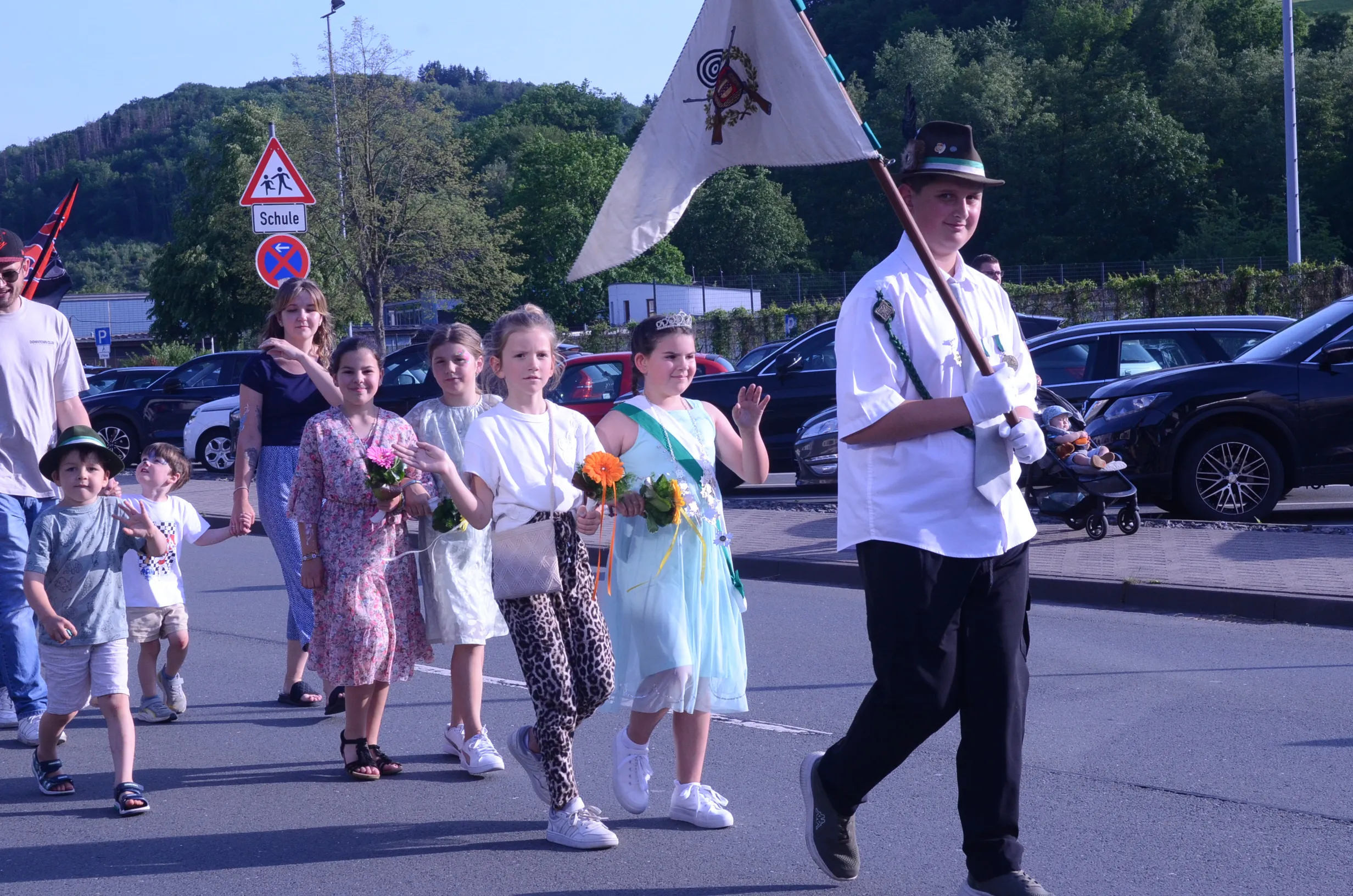 Ein Junge in Tracht führt eine Gruppe von Kindern und Erwachsenen durch eine Straße, während er eine Fahne trägt. Die Kinder halten Blumensträuße, und ein Mädchen trägt eine Krone.