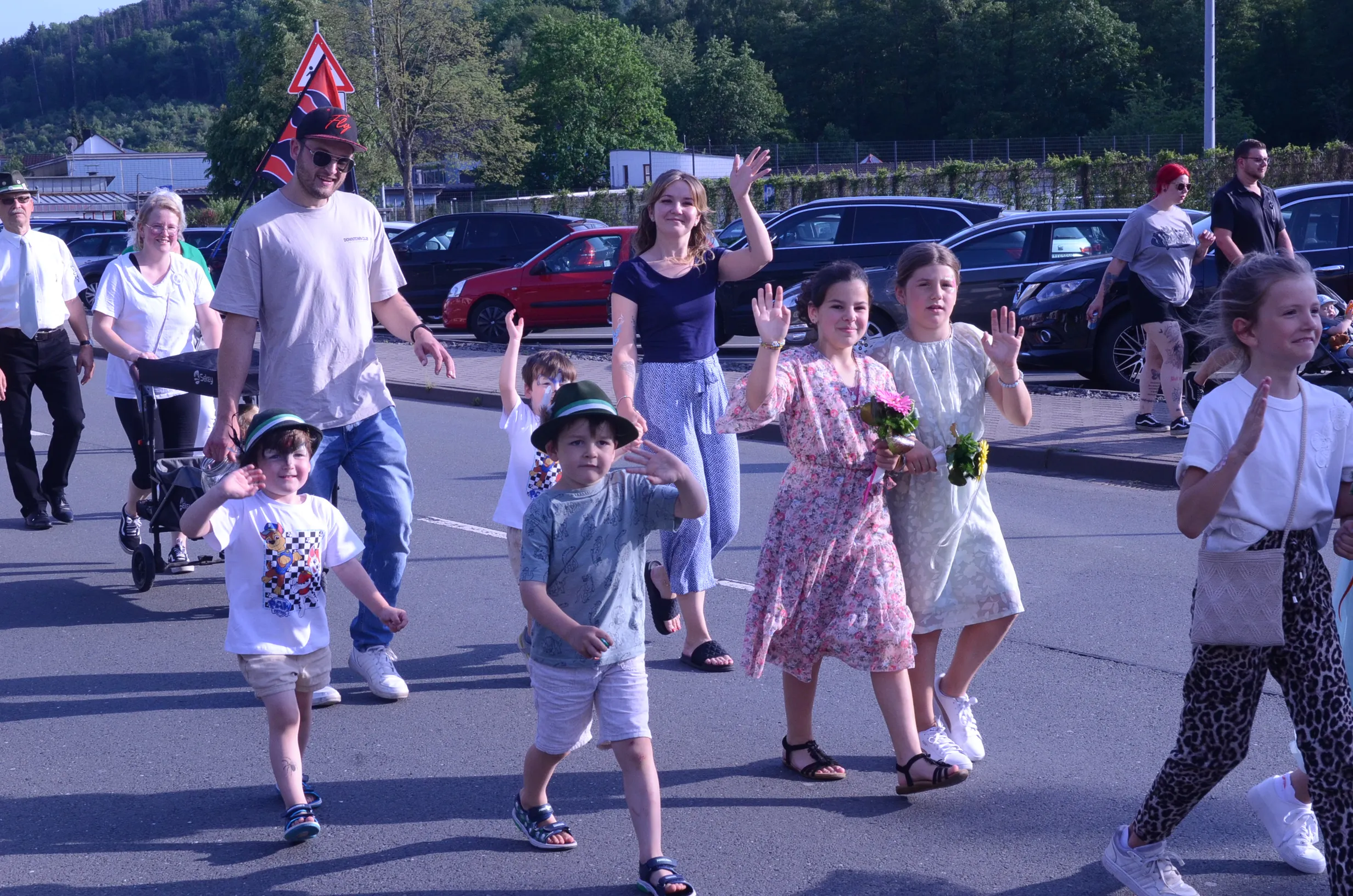 Eine Gruppe von Menschen, darunter Kinder und Erwachsene, winkt in einer Fußgängerzone. Im Hintergrund sind Autos und Bäume zu sehen.